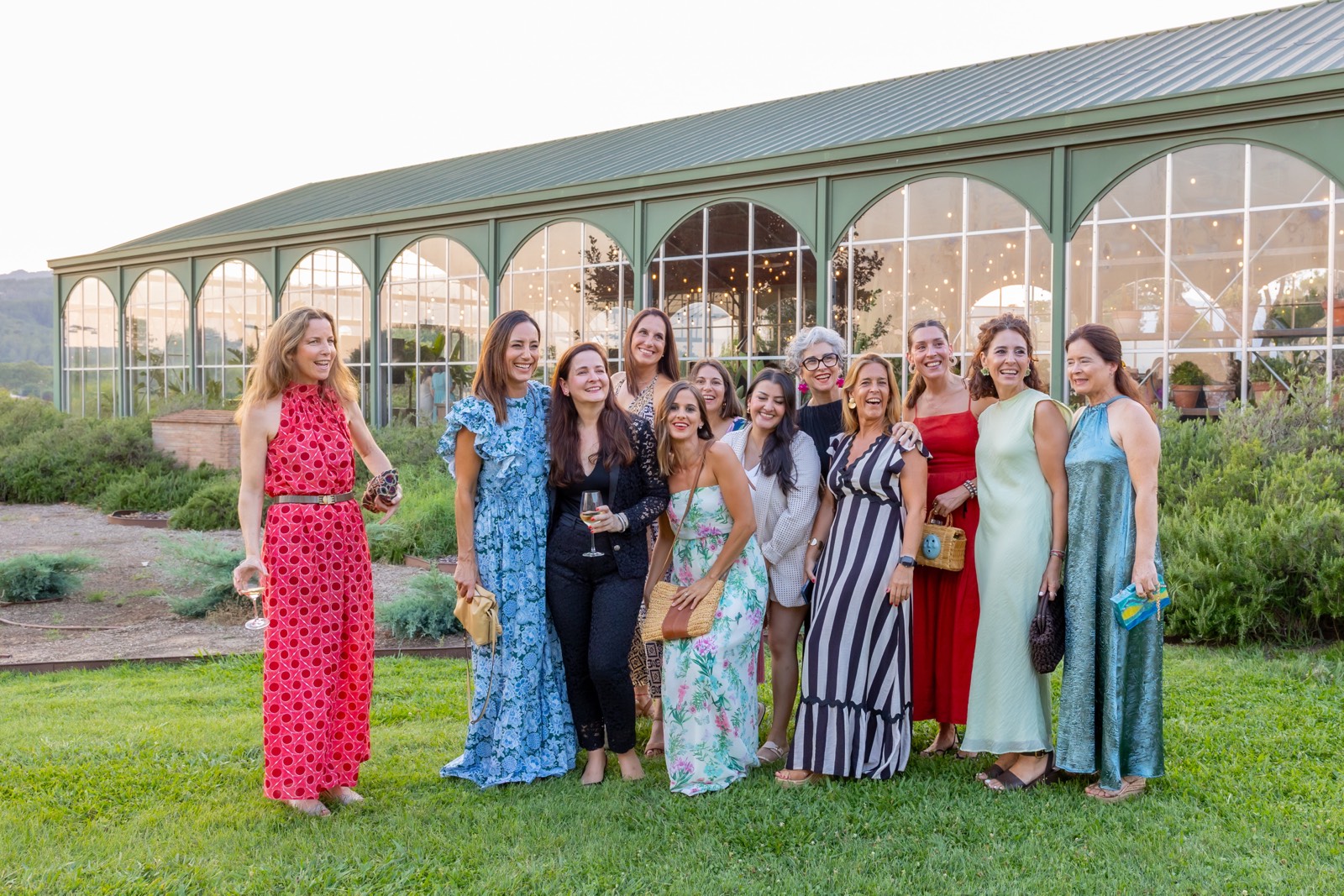 Grupo de mujeres vestidas elegantemente posando juntas frente a un edificio de cristal con arcos verdes, en un jardín con césped