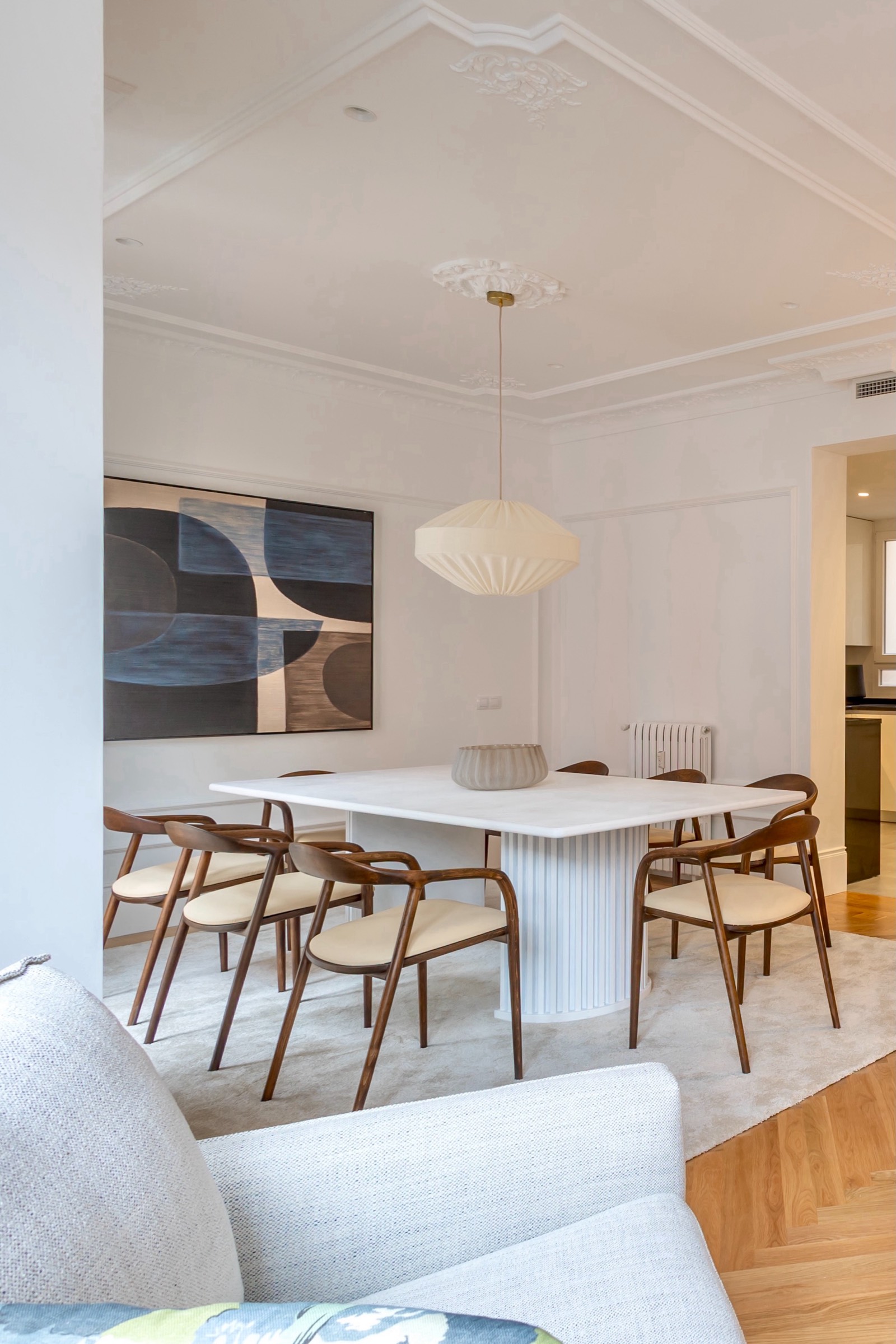 Comedor elegante con mesa blanca rectangular, sillas de madera con asientos claros, lámpara colgante moderna y cuadro abstracto en la pared. Decoración minimalista en tonos neutros con techo ornamentado.