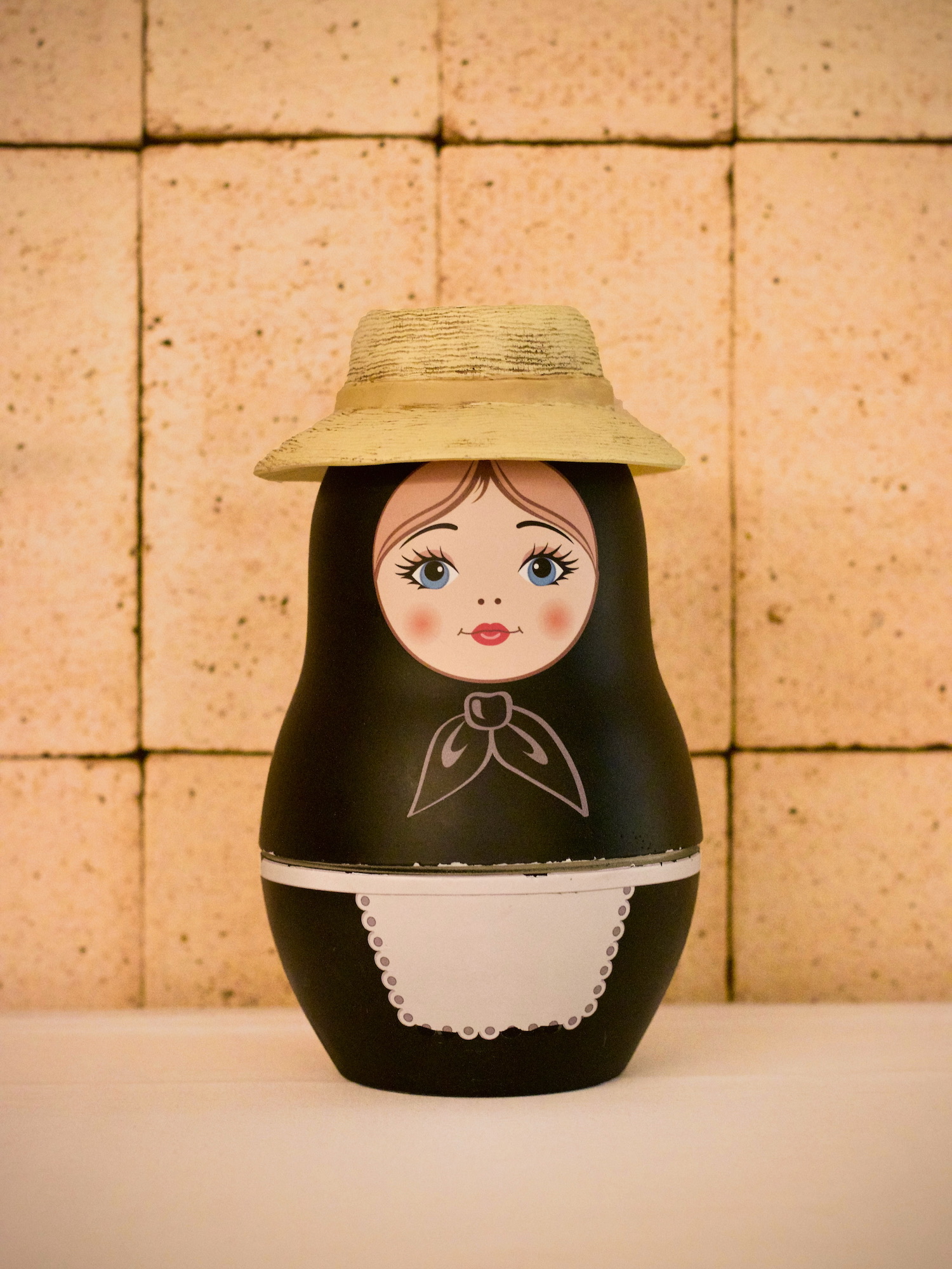 Muñeca matrioska decorativa negra con delantal blanco y sombrero de paja, exhibida sobre una superficie clara contra una pared con azulejos beige. La muñeca tiene detalles pintados tradicionales en su rostro y un pañuelo al cuello.
