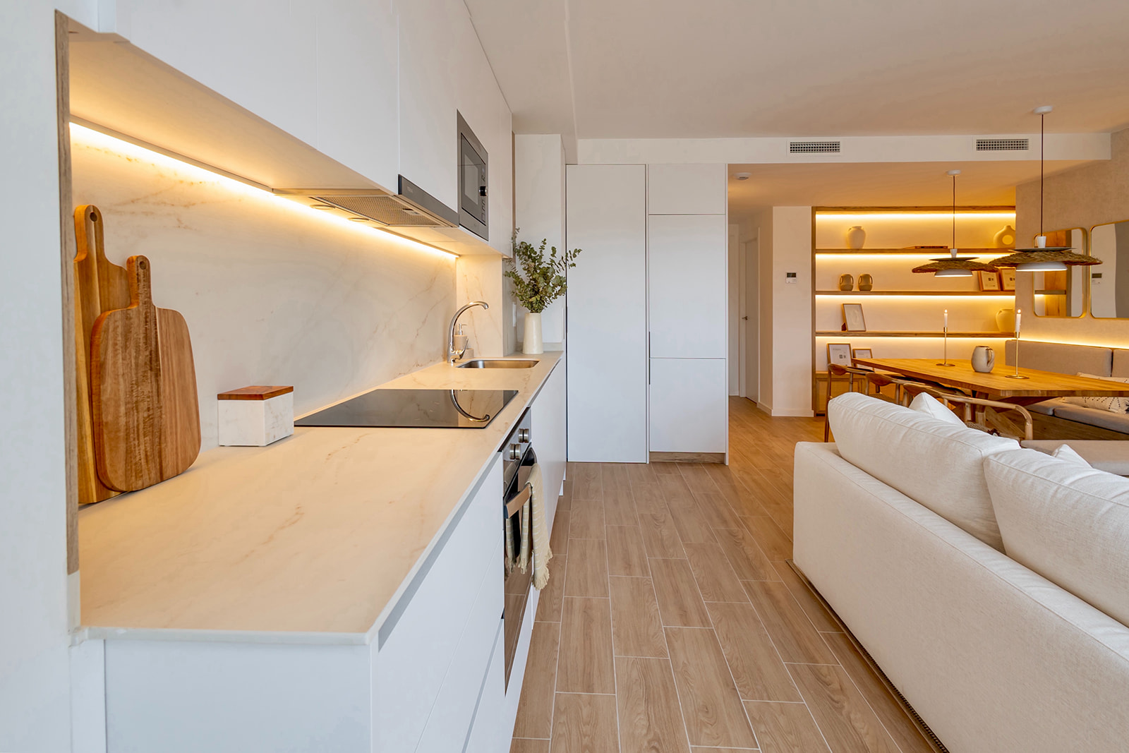 Cocina moderna abierta conectada con el salón. Encimera blanca con iluminación LED, electrodomésticos integrados y tablas de cortar de madera. Suelo de madera y sofá blanco en primer plano. Iluminación cálida y ambiente acogedor.