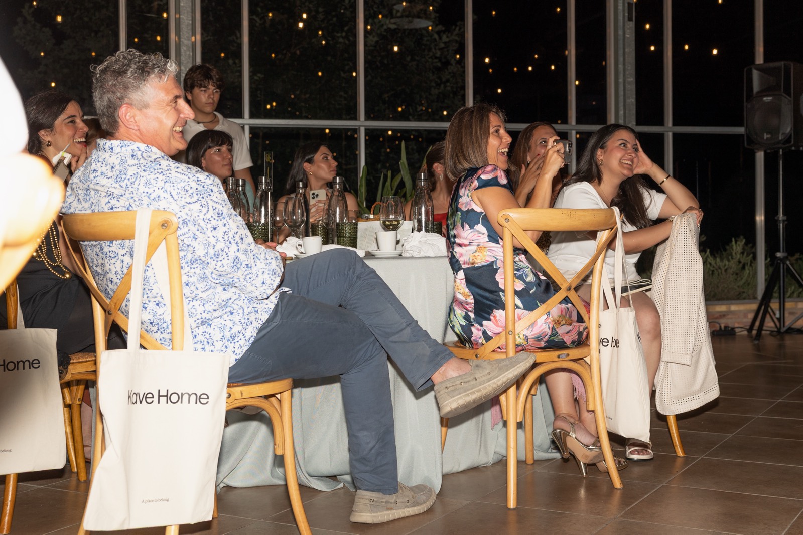 Grupo de personas sentadas en sillas de madera en un evento nocturno con iluminación decorativa, riendo y socializando
