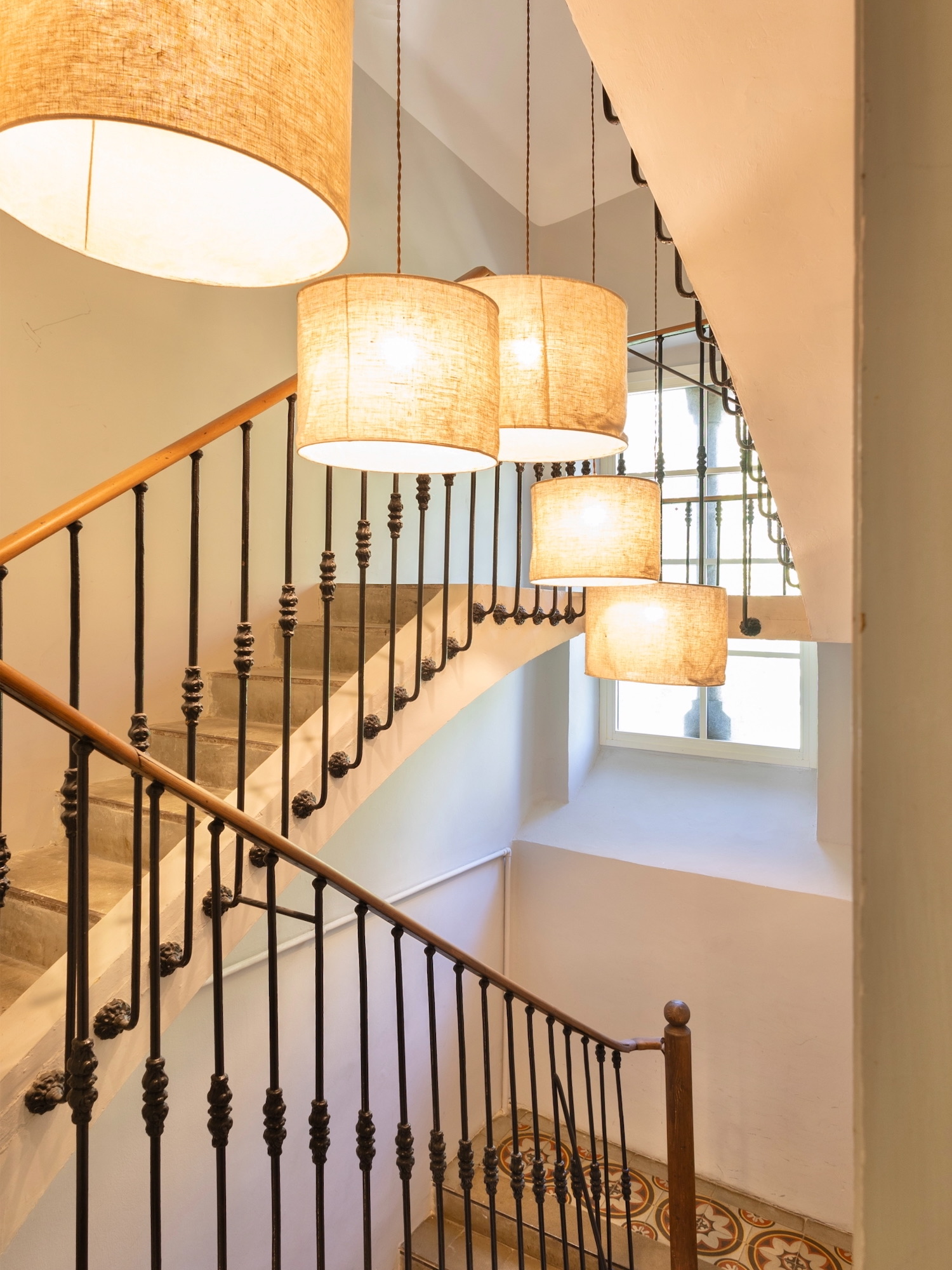 Elegante escalera curva con barandilla de hierro forjado decorativo, iluminada por una serie de lámparas colgantes cilíndricas con pantallas beige. El diseño combina elementos clásicos y modernos en un espacio luminoso.
