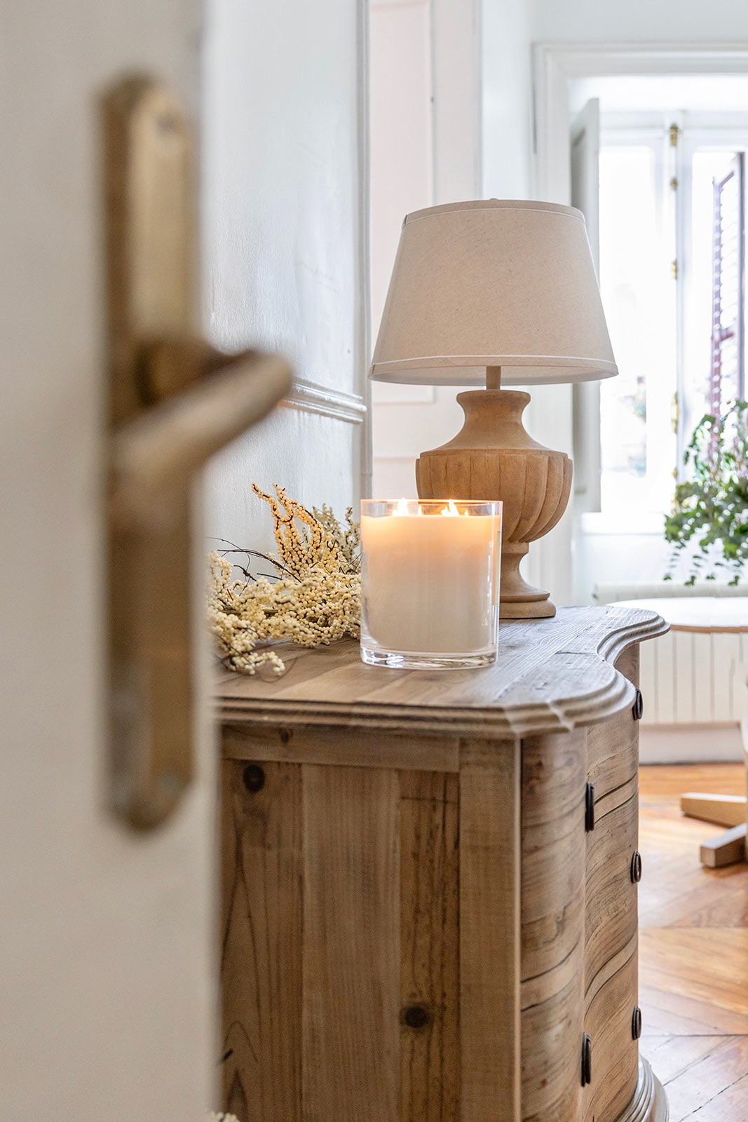 Decoración hogareña con estilo rústico que incluye una cómoda de madera clara, una lámpara con base de madera y pantalla beige, una vela aromática en vaso de cristal y flores secas decorativas