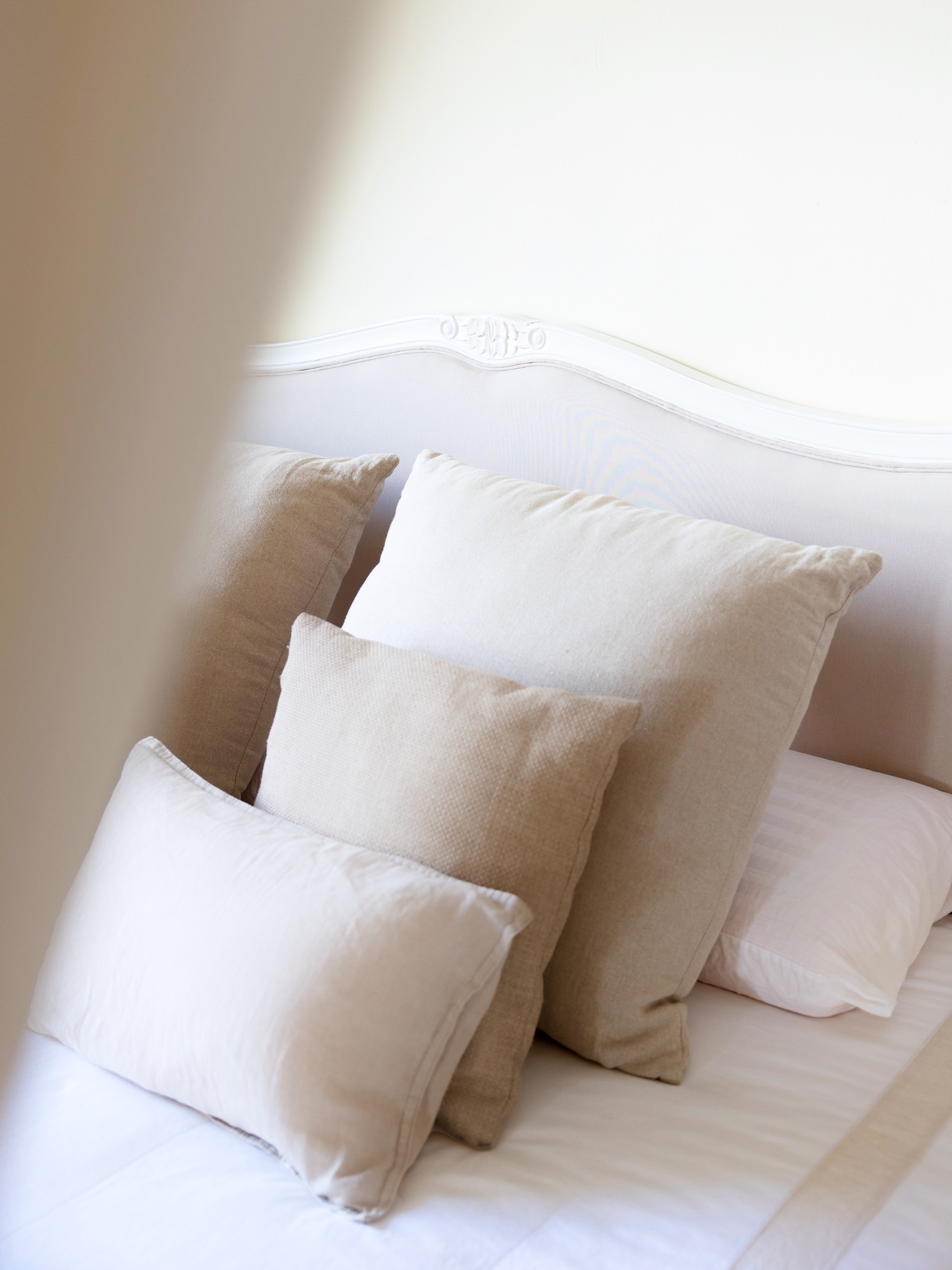 Elegante composición de cama con almohadas en tonos blancos y beige apoyadas sobre un respaldo de cama vintage blanco con detalles decorativos tallados