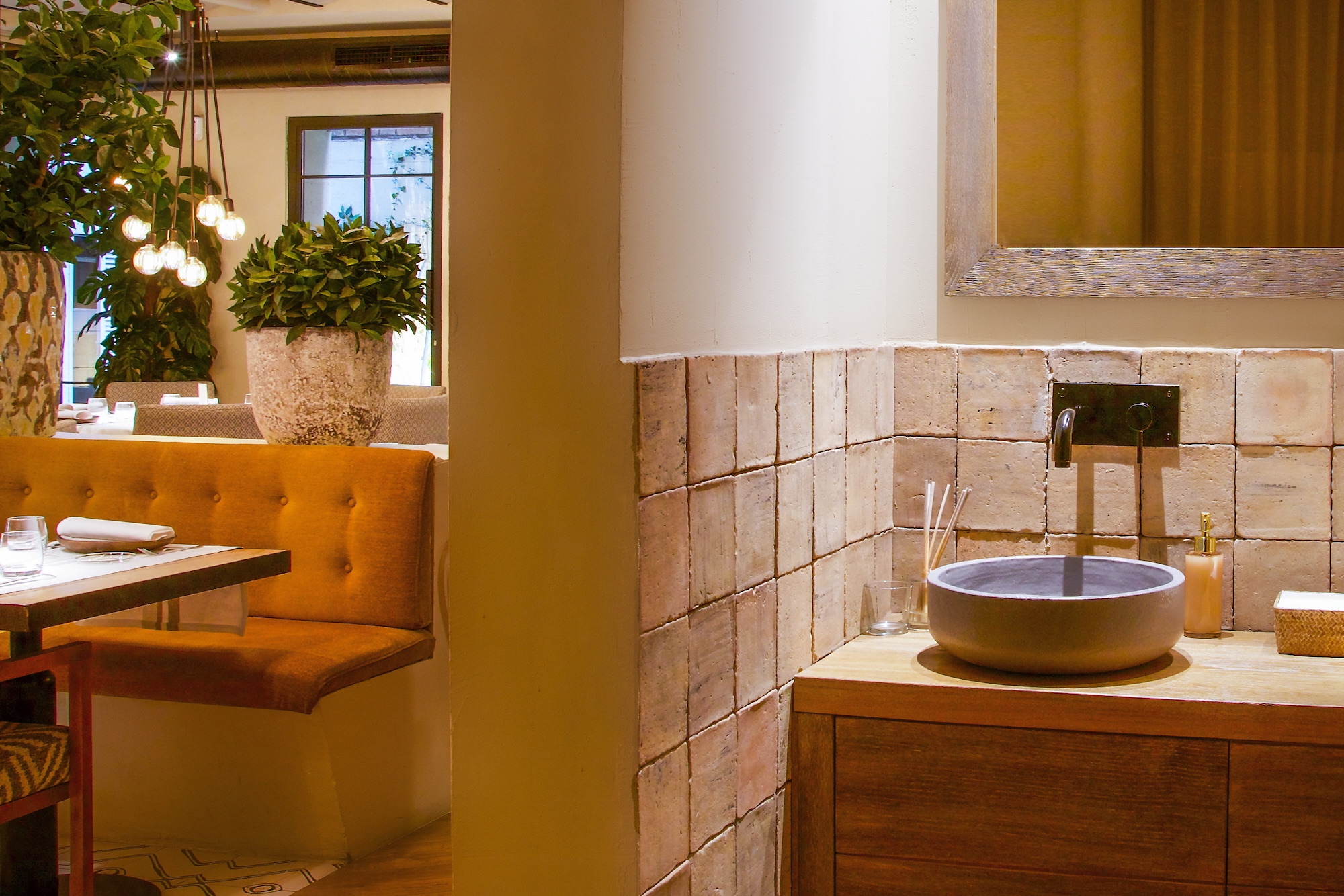 Interior de restaurante elegante con decoración rústica moderna, que muestra un lavabo circular gris sobre mueble de madera, pared de azulejos claros, asientos tapizados en naranja y plantas decorativas con iluminación colgante