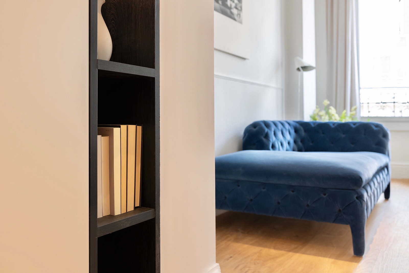 Interior de un salón moderno con un sofá azul de terciopelo acolchado y una estantería negra con libros. La habitación tiene paredes blancas, piso de madera y una ventana con cortinas blancas.