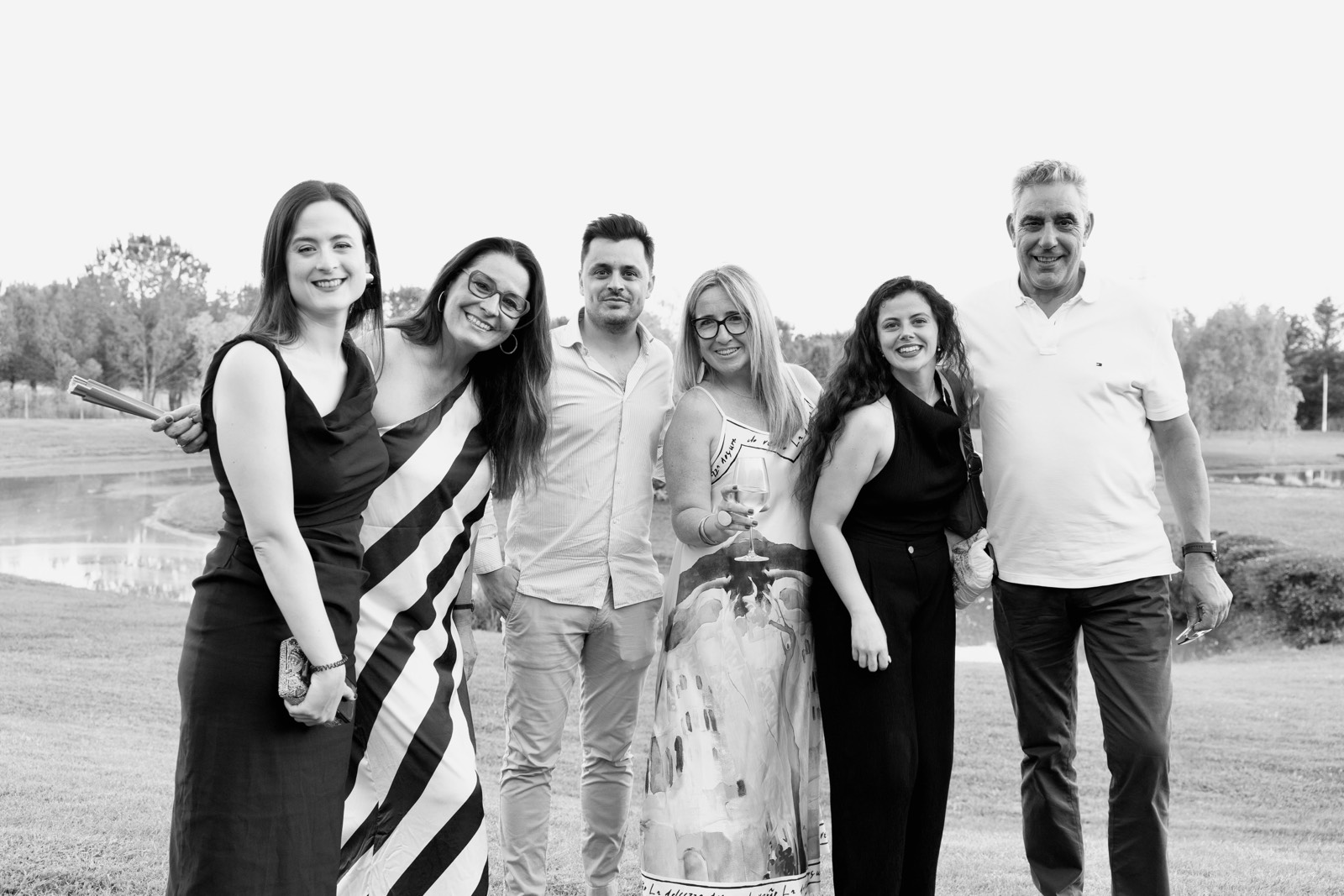 Grupo de seis personas sonrientes posando juntas al aire libre en un entorno de césped, vestidos con ropa elegante en blanco y negro