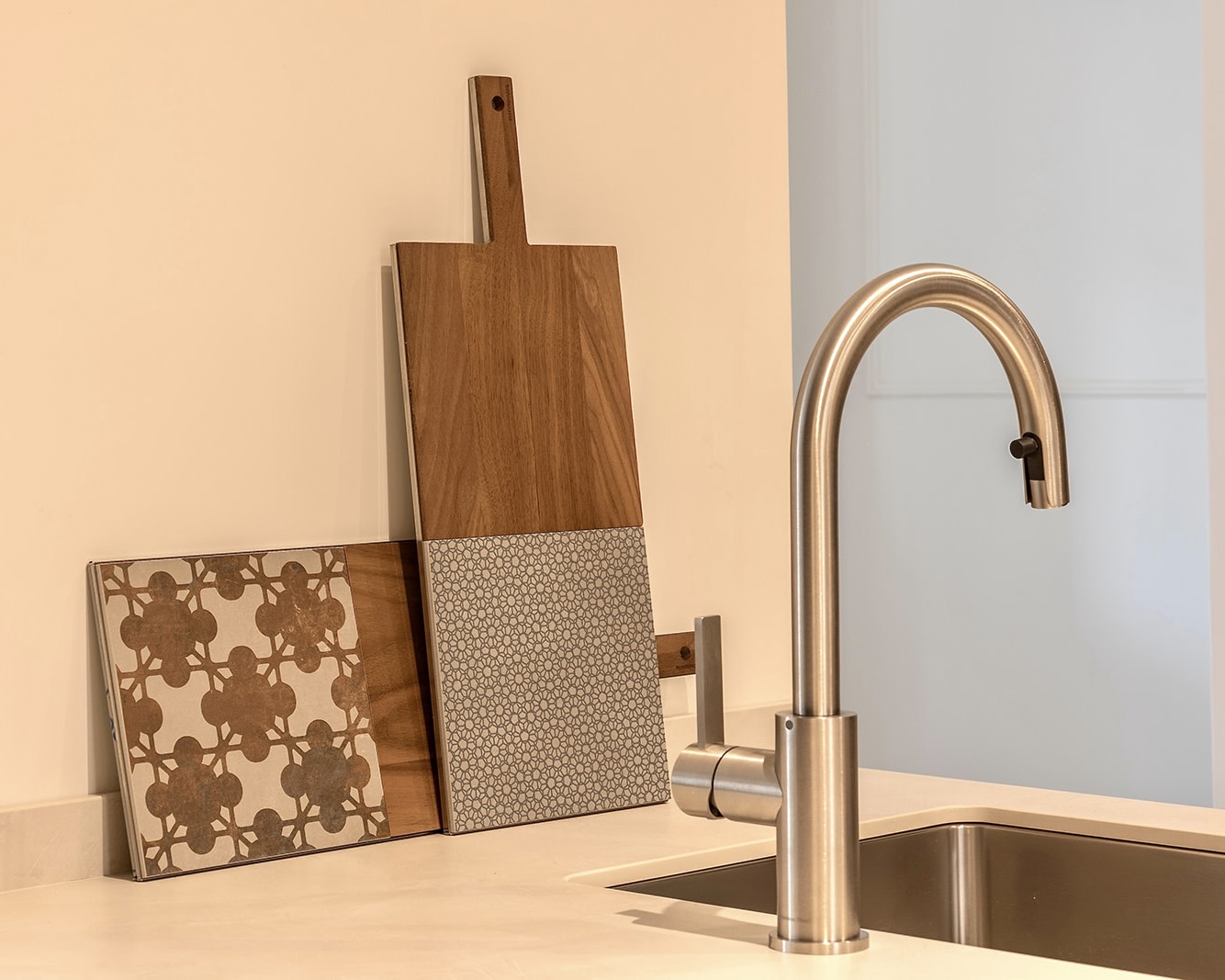 Cocina moderna con fregadero de acero inoxidable y grifo de níquel cepillado. Una tabla de cortar de madera y azulejos decorativos se apoyan contra la pared. Los colores predominantes son beige y marrón claro.