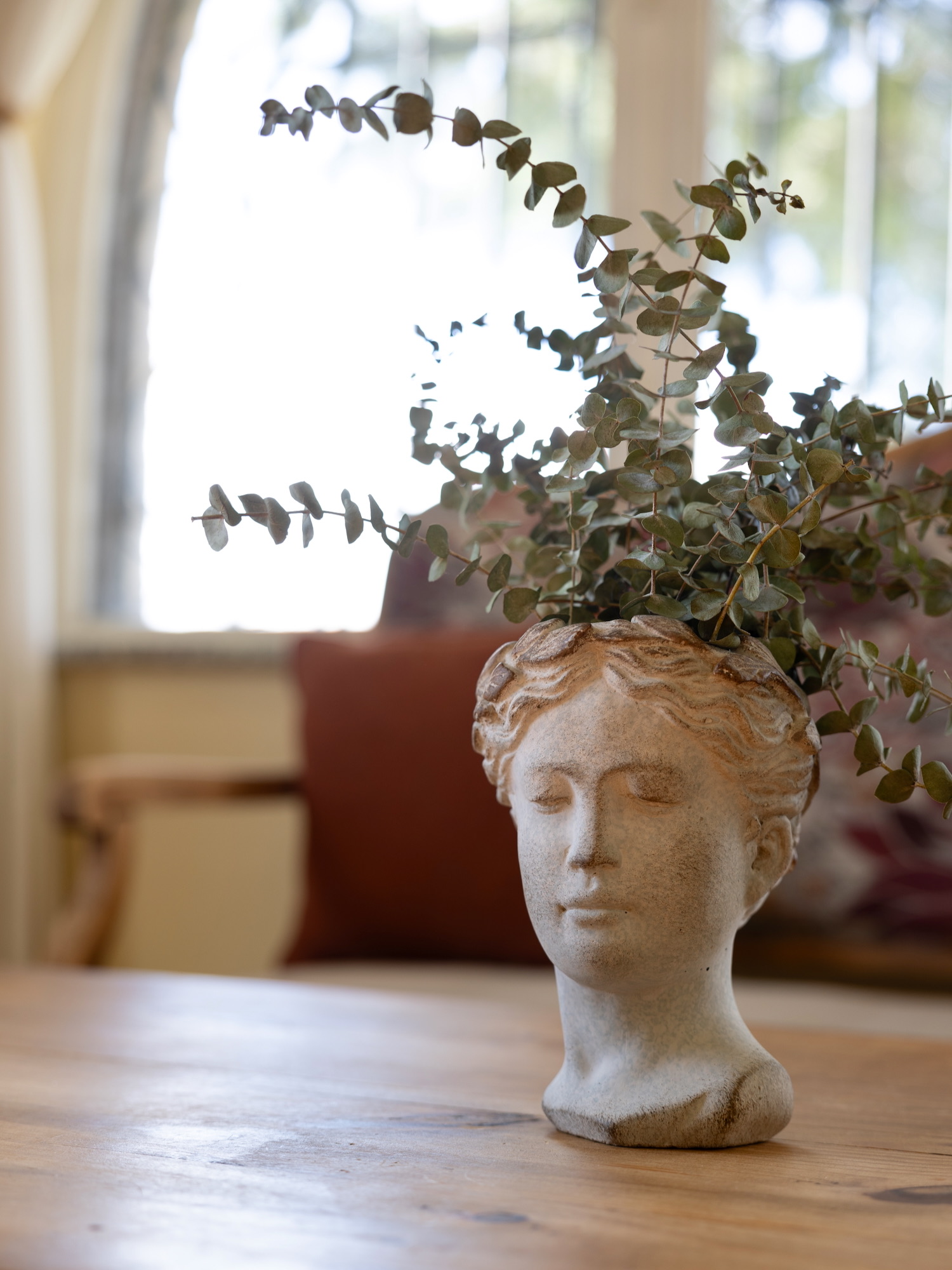 Busto decorativo estilo griego en color blanco con hojas de eucalipto que sobresalen de la parte superior, colocado sobre una mesa de madera con un fondo desenfocado y suave