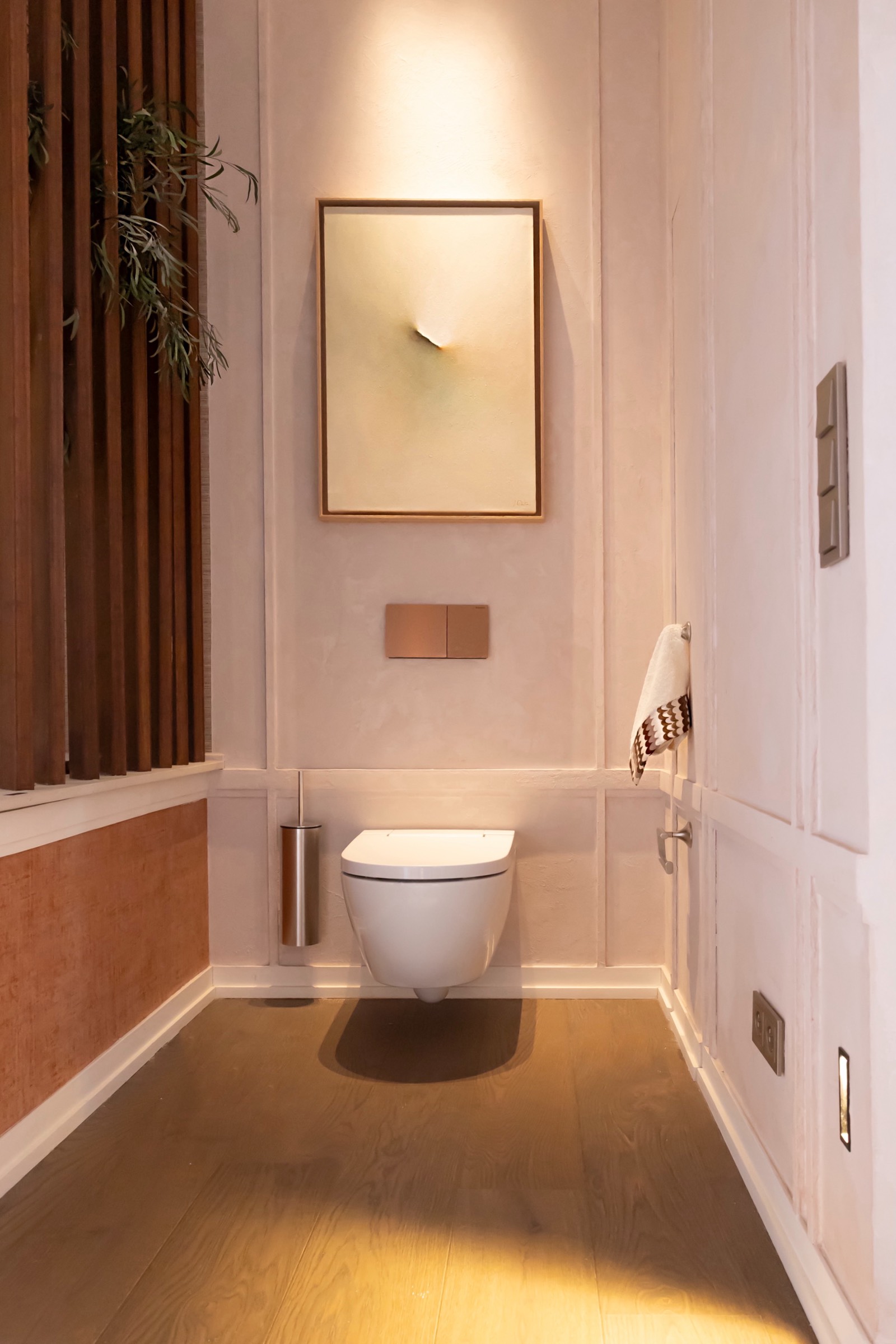 Baño contemporáneo con inodoro suspendido blanco, paredes neutras, piso de madera y decoración minimalista con planta y arte en la pared