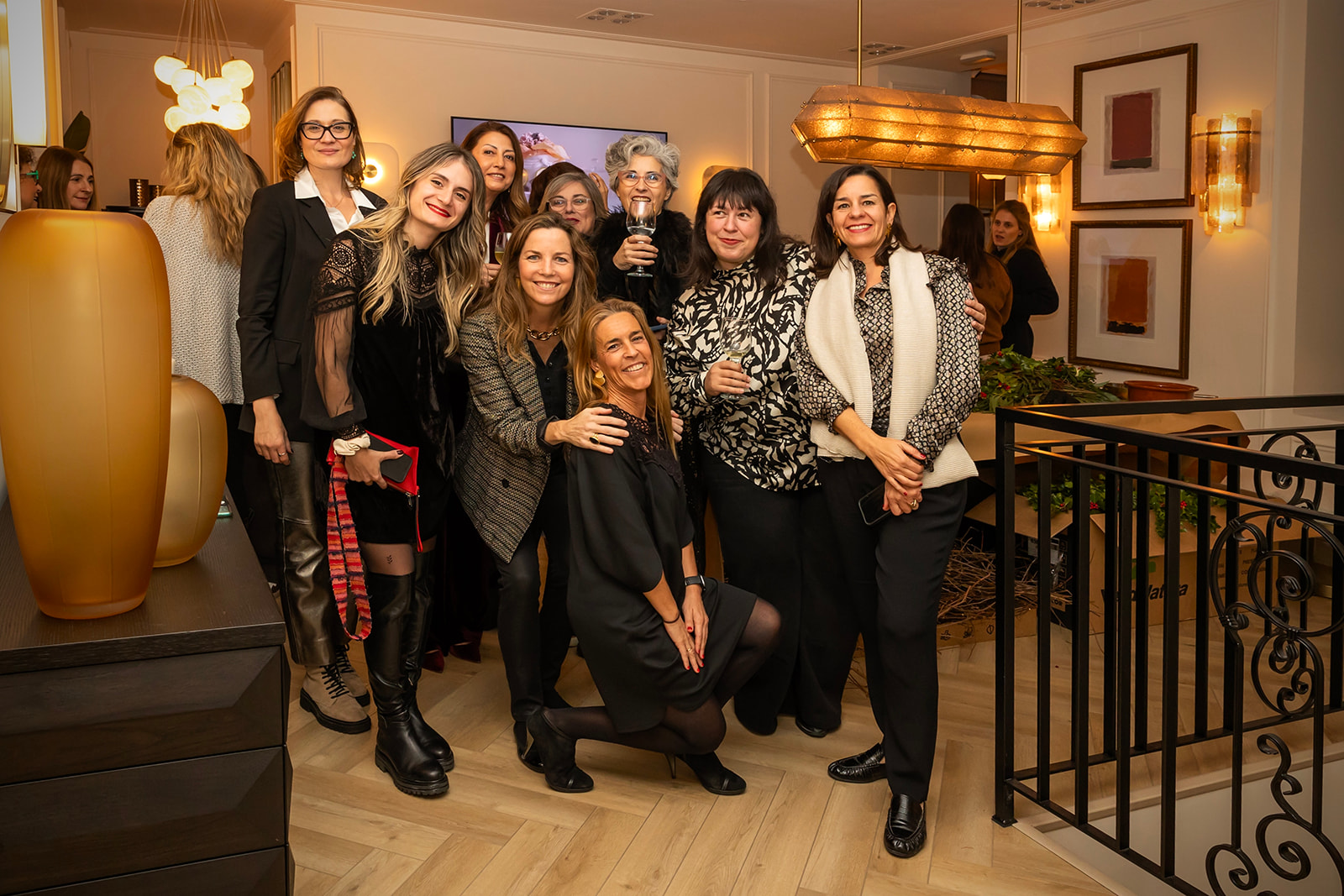 Grupo de mujeres sonrientes posando juntas en una elegante sala de estar con iluminación cálida. Están vestidas con ropa casual y formal, creando una atmósfera de celebración o reunión social.