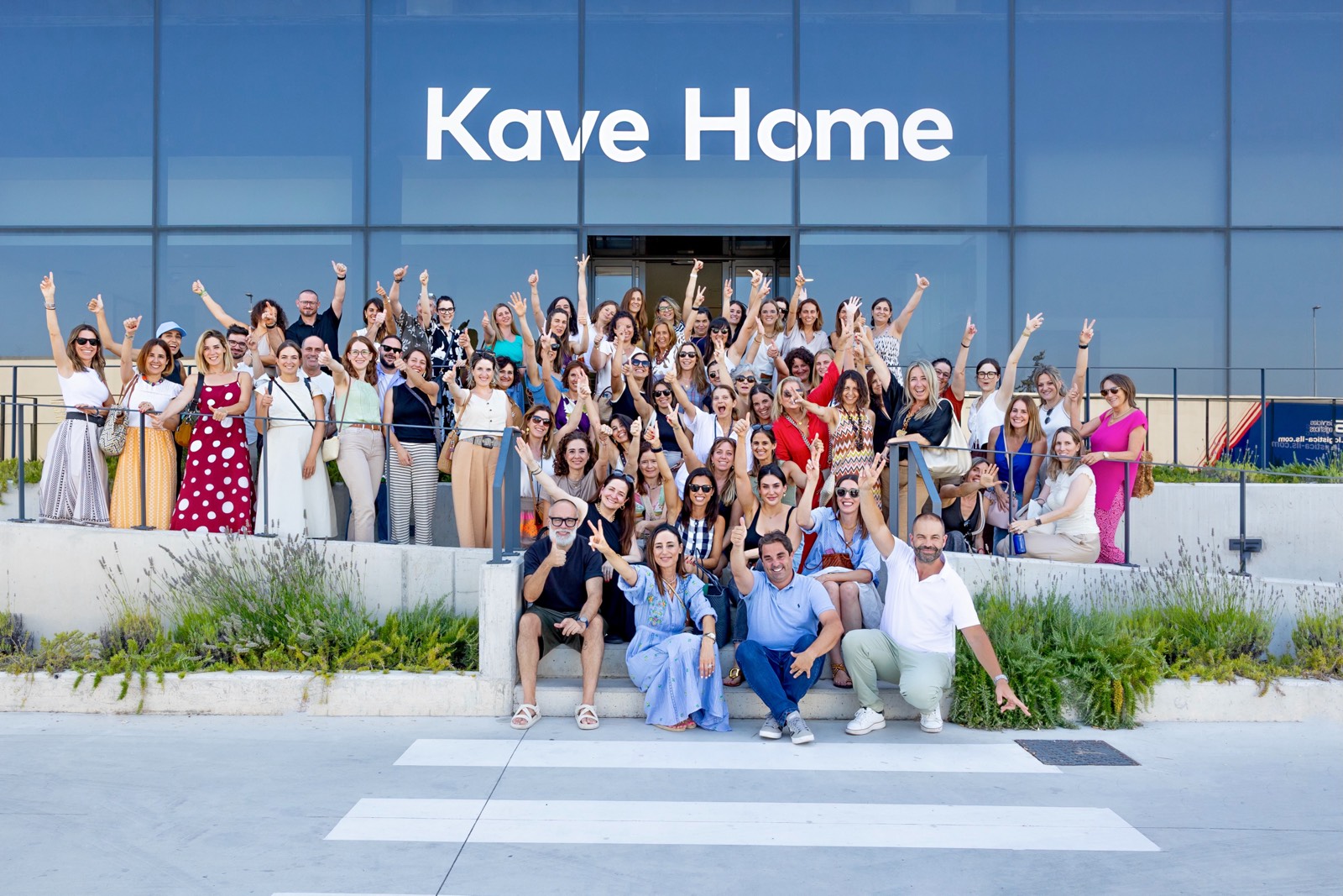 Gran grupo de empleados de Kave Home posando frente a su edificio corporativo, levantando los brazos con entusiasmo y alegría