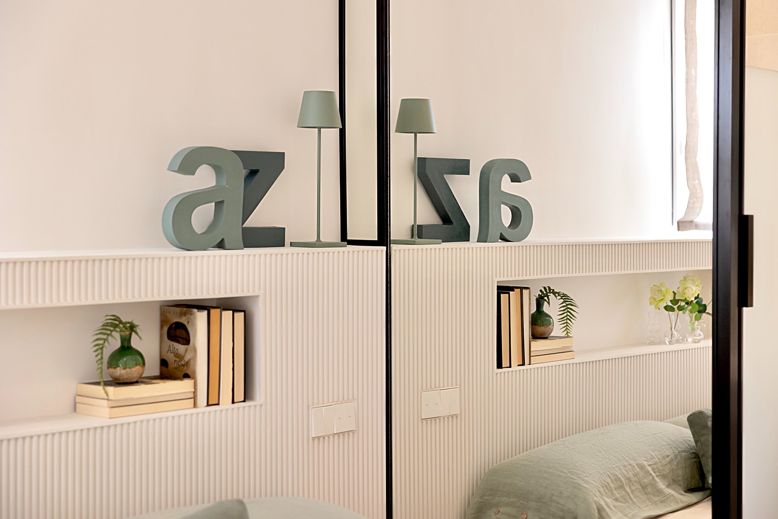 Decoración minimalista de una habitación con letras grandes 'az' en verde sage, lámparas pequeñas y estanterías empotradas con libros y plantas. Los colores predominantes son el beige y el verde suave.