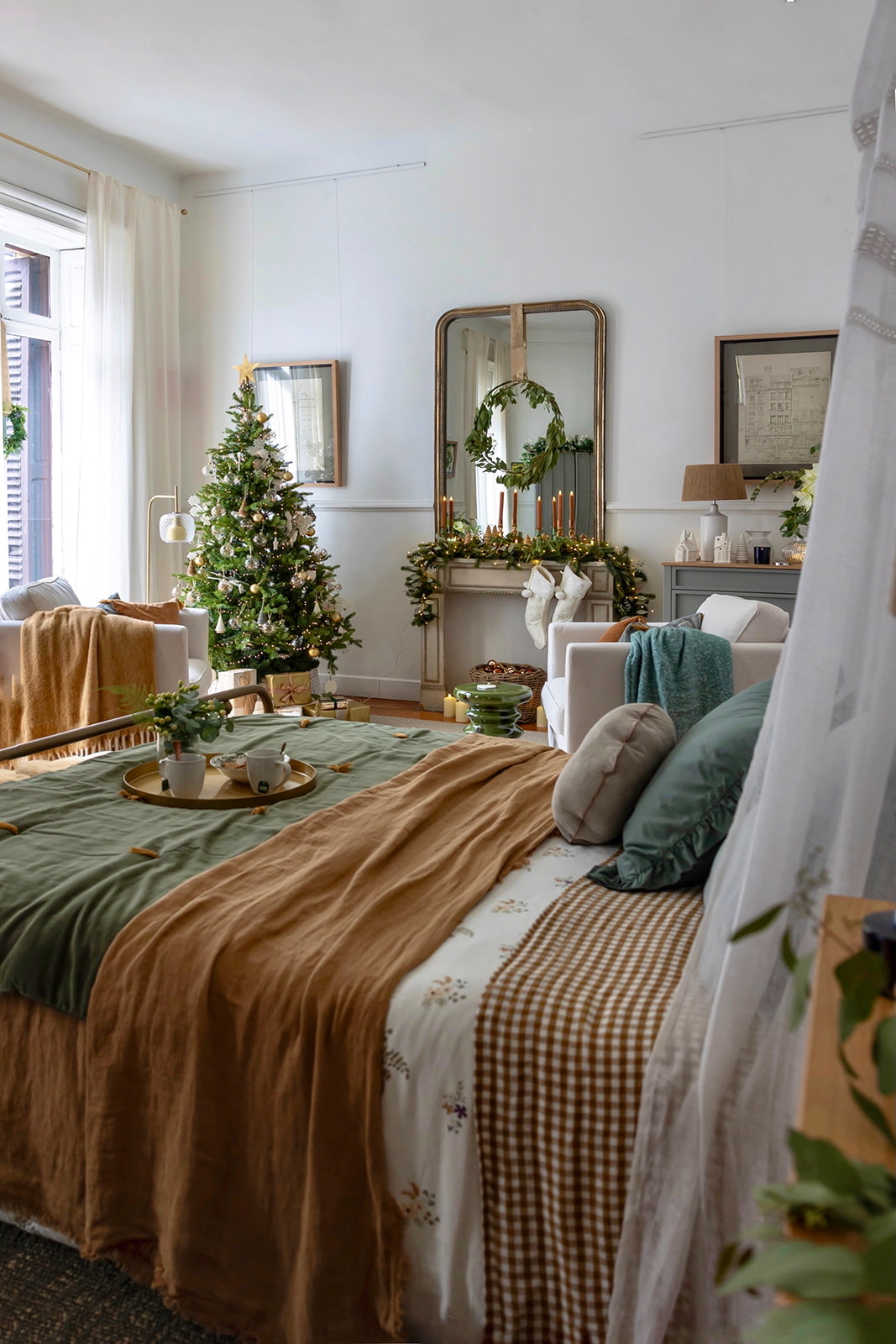 Dormitorio decorado para Navidad con árbol adornado, guirnaldas verdes, espejo dorado y ropa de cama en tonos tierra y verde. Estilo rústico elegante con detalles festivos como calcetines navideños y velas.
