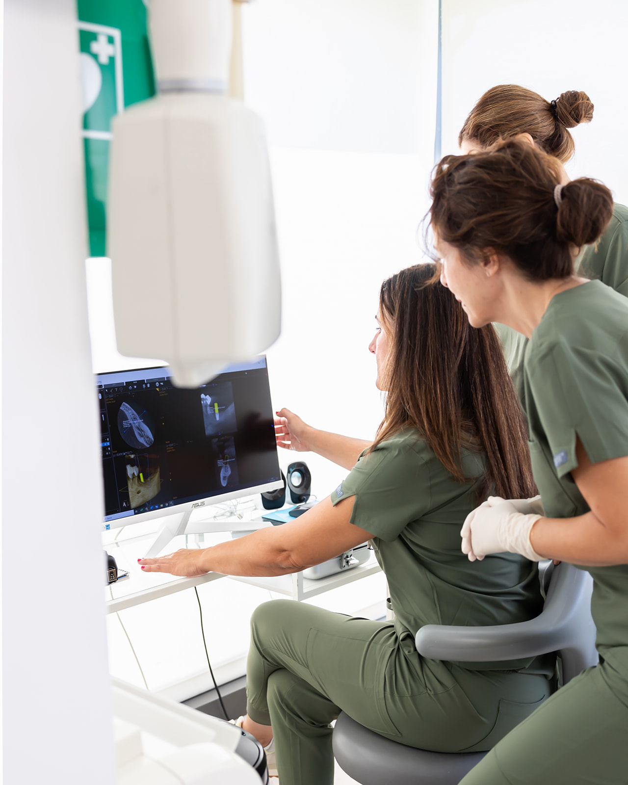 Profesionales médicos en uniformes verdes observan imágenes diagnósticas en una pantalla de computadora en un entorno clínico. Se aprecia un equipo médico y un símbolo de cruz en el fondo.