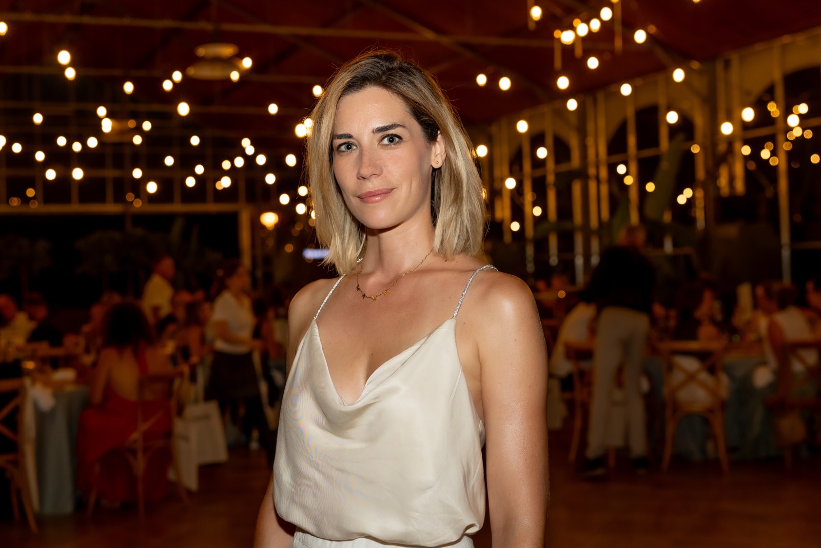 Persona en vestido blanco de tirantes en un evento nocturno con iluminación cálida de guirnaldas, ambiente elegante con mesas y personas en el fondo