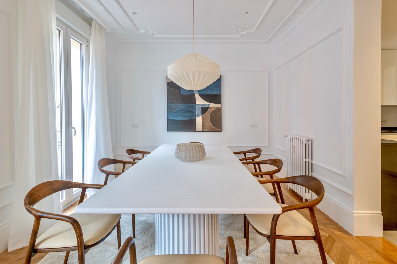 Comedor elegante con mesa blanca rectangular, sillas de madera curvas, lámpara colgante blanca y cuadro abstracto en la pared. La habitación tiene paredes blancas, molduras decorativas y ventanas grandes con cortinas claras.