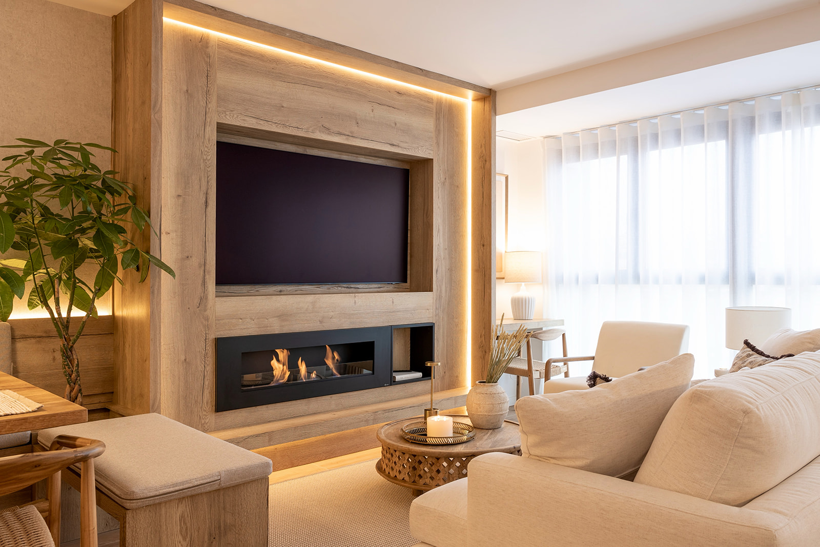 Sala de estar moderna con chimenea integrada y televisión de pantalla plana sobre un marco de madera iluminado. Muebles en tonos claros y una planta decorativa crean un ambiente acogedor y luminoso.