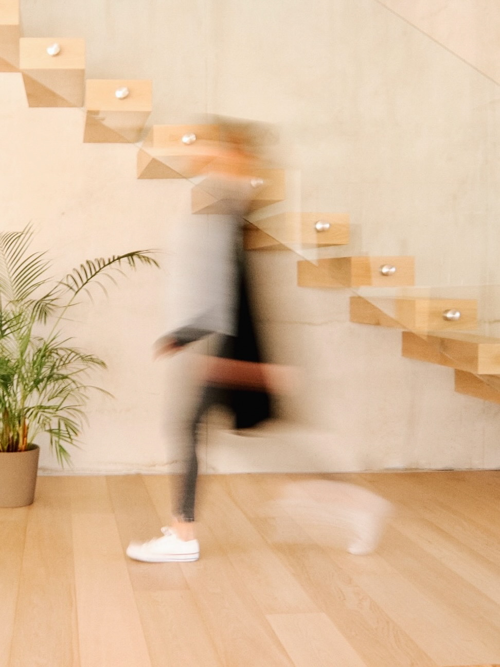 Imagen borrosa de movimiento que muestra una silueta subiendo unas escaleras flotantes de madera con iluminación LED. Una planta tropical decora la esquina inferior, sobre un piso de madera clara.