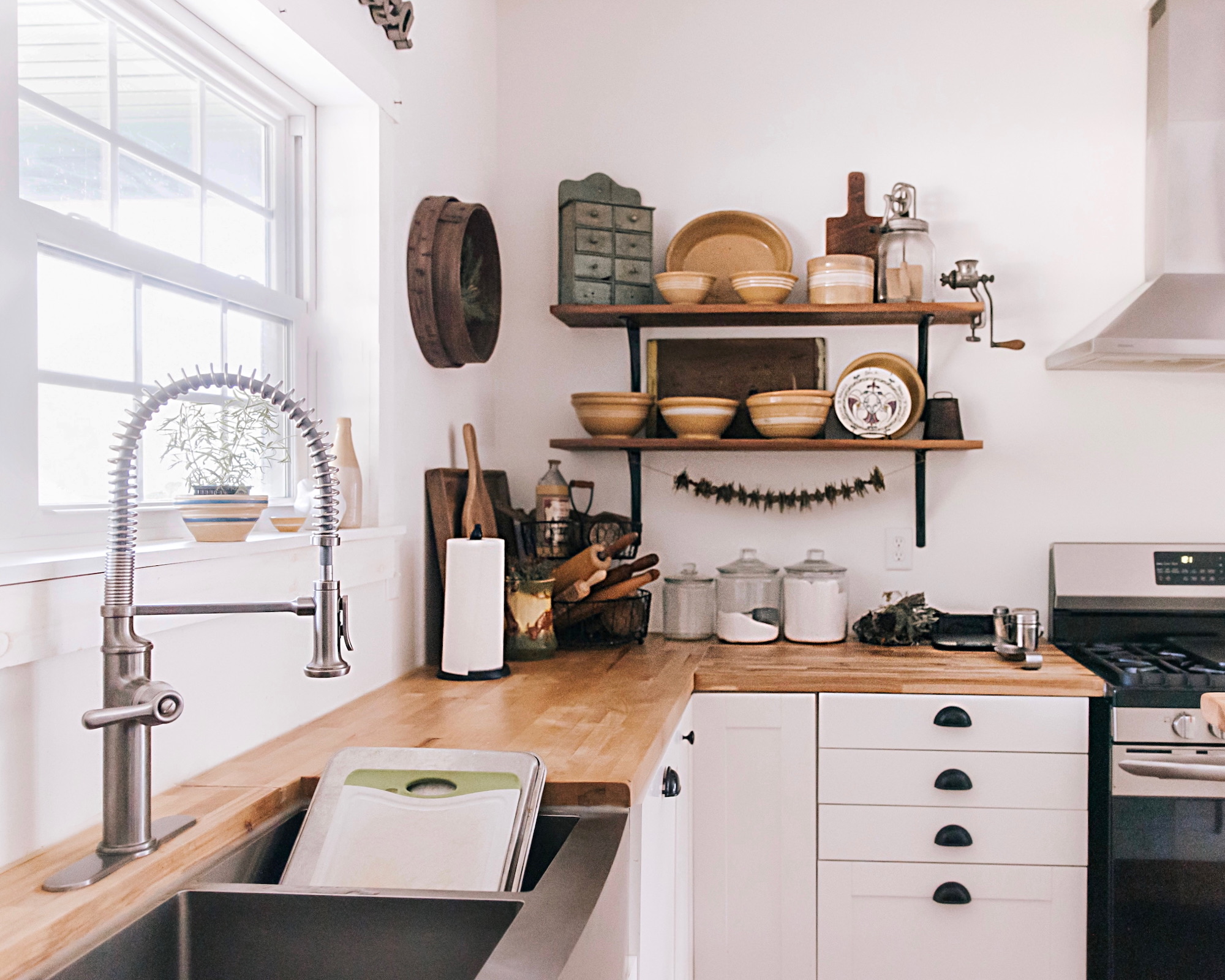 Cocina moderna con encimeras de madera, gabinetes blancos y estantes rústicos decorados con cuencos de cerámica y accesorios vintage. Cuenta con un grifo profesional de acero y una ventana que aporta luz natural.