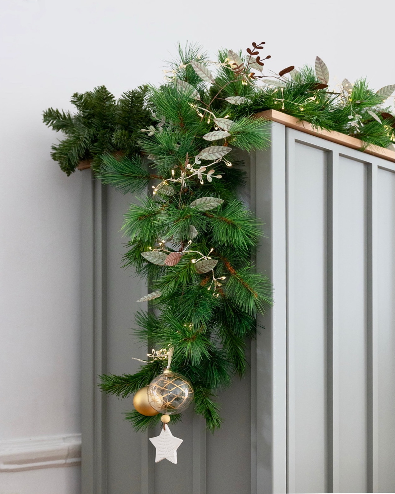 Guirnalda navideña de pino artificial decorada con hojas plateadas, luces LED y adornos dorados incluyendo una esfera y una estrella blanca, colocada sobre un mueble gris