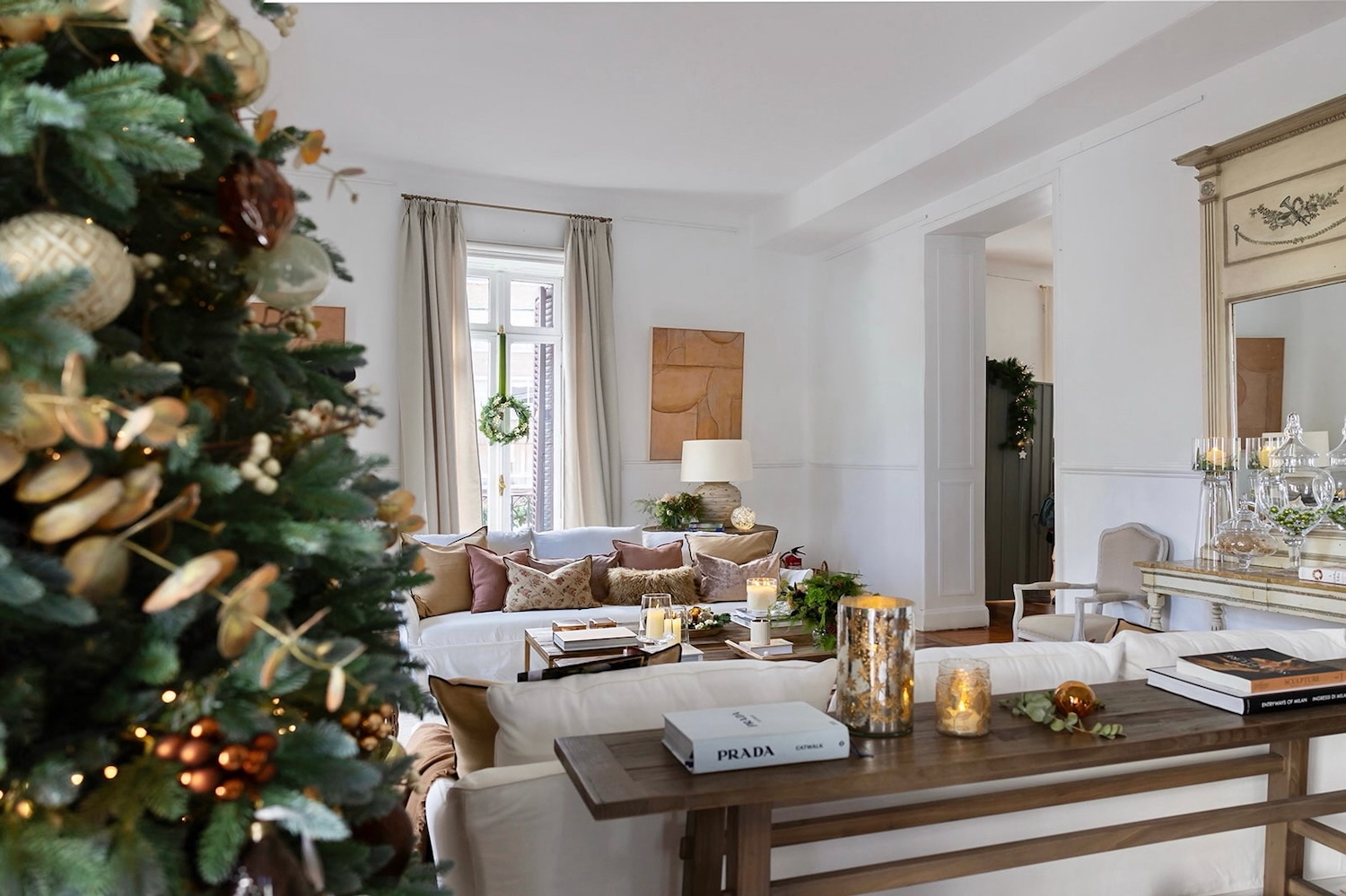 Sala de estar elegante decorada para Navidad con árbol adornado en tonos dorados, sofás blancos, mesa de consola de madera, velas decorativas y detalles festivos en un ambiente luminoso y moderno