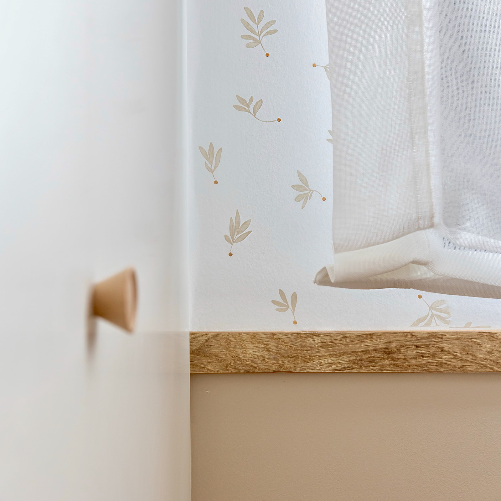 Cortina blanca translúcida con estampado de hojas beige, colgada sobre un alféizar de madera. Se aprecia un tirador de puerta desenfocado en primer plano, creando una atmósfera luminosa y minimalista.