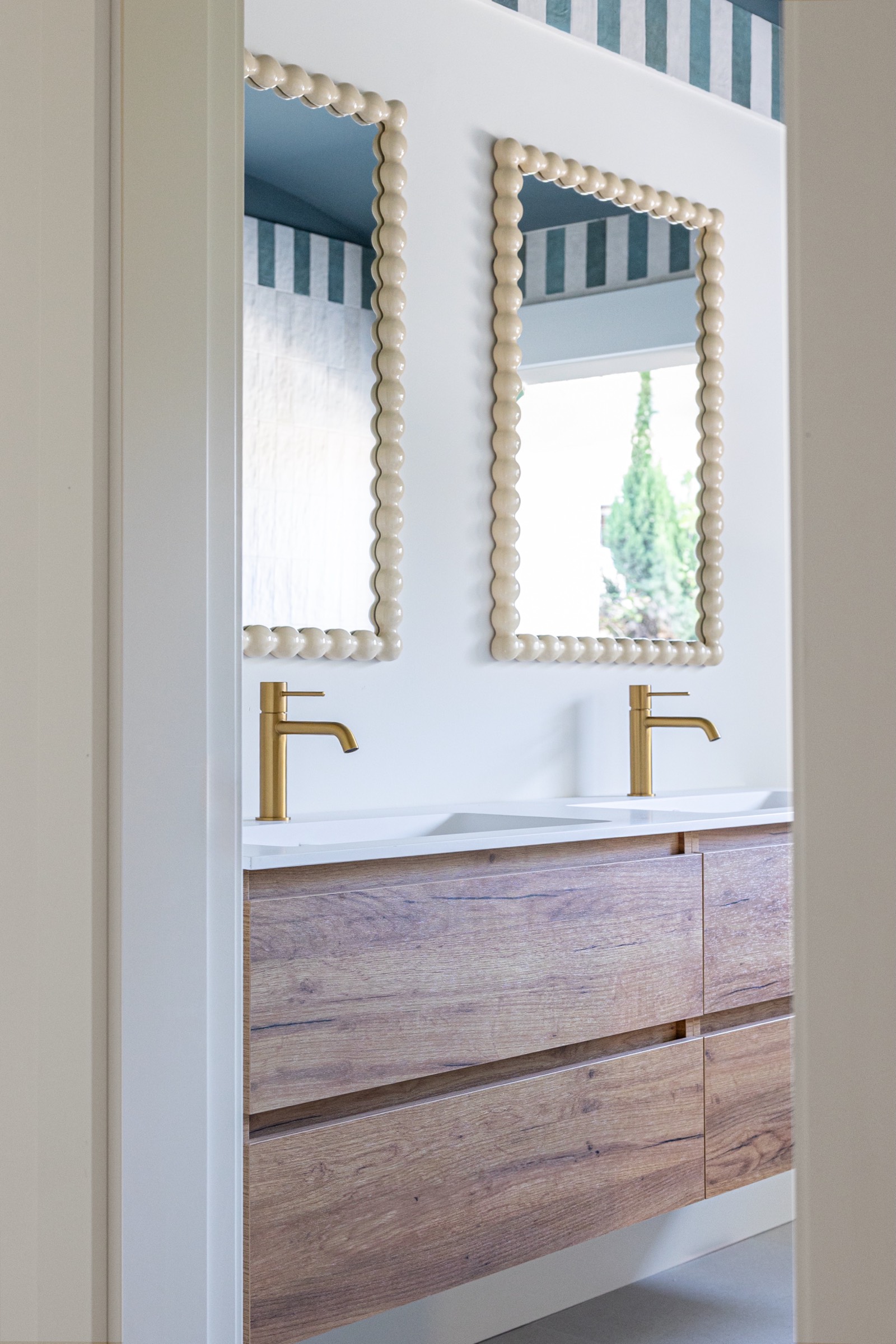 Detalle de un lavabo de baño moderno con mueble de madera, dos espejos con marco ondulado y grifos dorados sobre encimera blanca