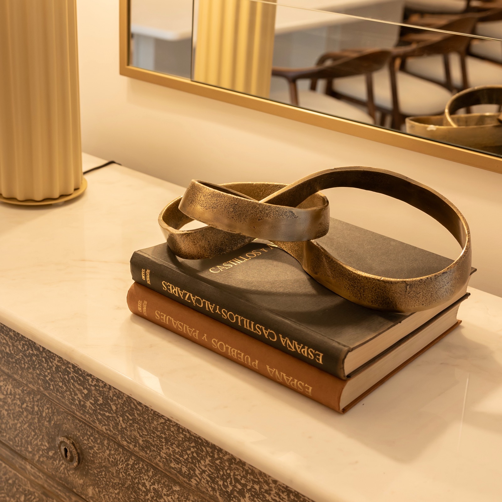 Imagen de una mesa con dos libros apilados y un adorno dorado en forma de cinta encima. Se ve un espejo y una columna dorada en el fondo, creando una atmósfera elegante y sofisticada.