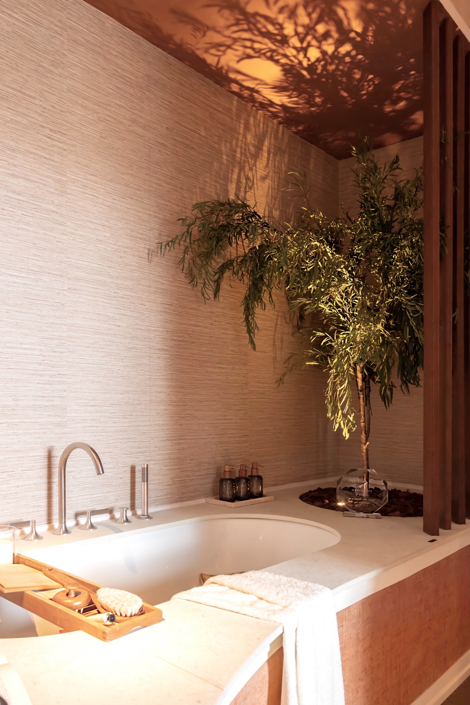 Bañera blanca con accesorios de madera, planta decorativa y sombras cálidas proyectadas en la pared con textura
