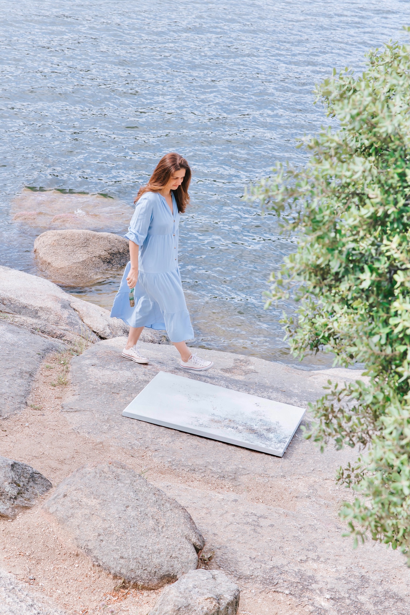 Mujer caminando por una costa rocosa junto al mar, vistiendo un vestido azul claro y zapatillas blancas. El paisaje incluye rocas, vegetación y agua azul.