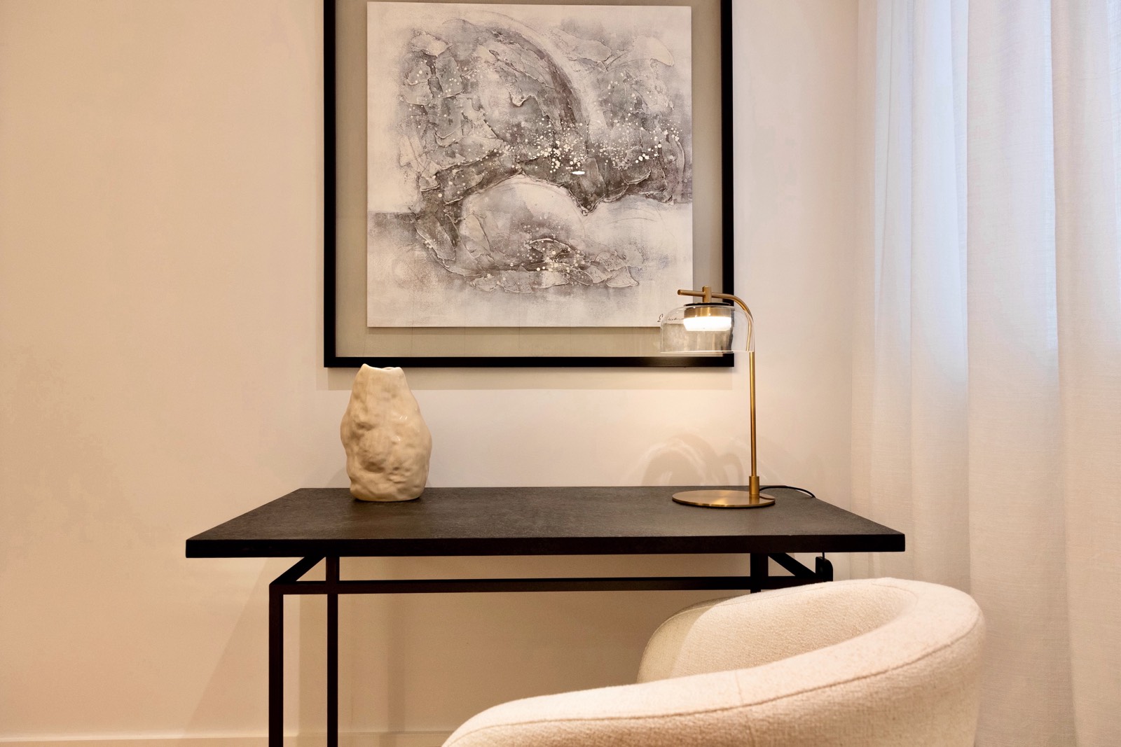 Rincón elegante con escritorio negro, silla beige, lámpara dorada y cuadro abstracto en tonos grises. La decoración incluye un jarrón blanco sobre el escritorio y cortinas blancas que aportan luminosidad al espacio.