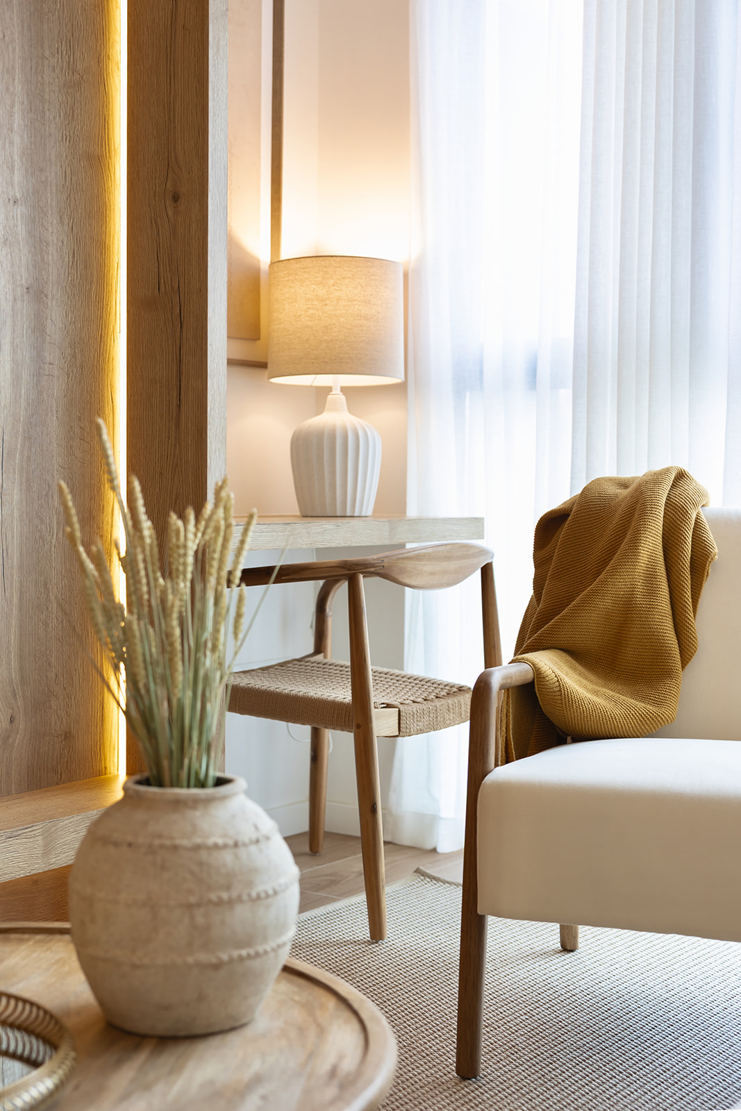 Un rincón acogedor con decoración natural que incluye una silla con manta amarilla, una lámpara blanca sobre una mesa auxiliar, cortinas blancas y un jarrón con plantas secas en primer plano.