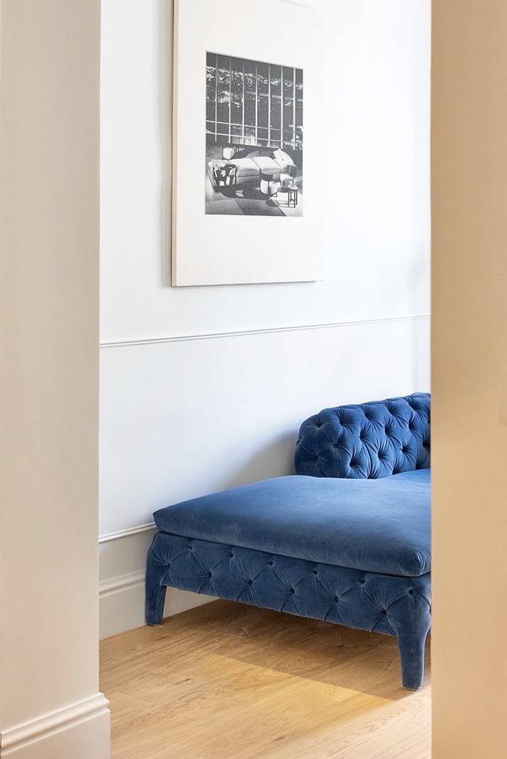 Sofá azul de terciopelo con botones en una habitación blanca con suelo de madera. Sobre el sofá hay un cuadro en blanco y negro enmarcado en la pared.