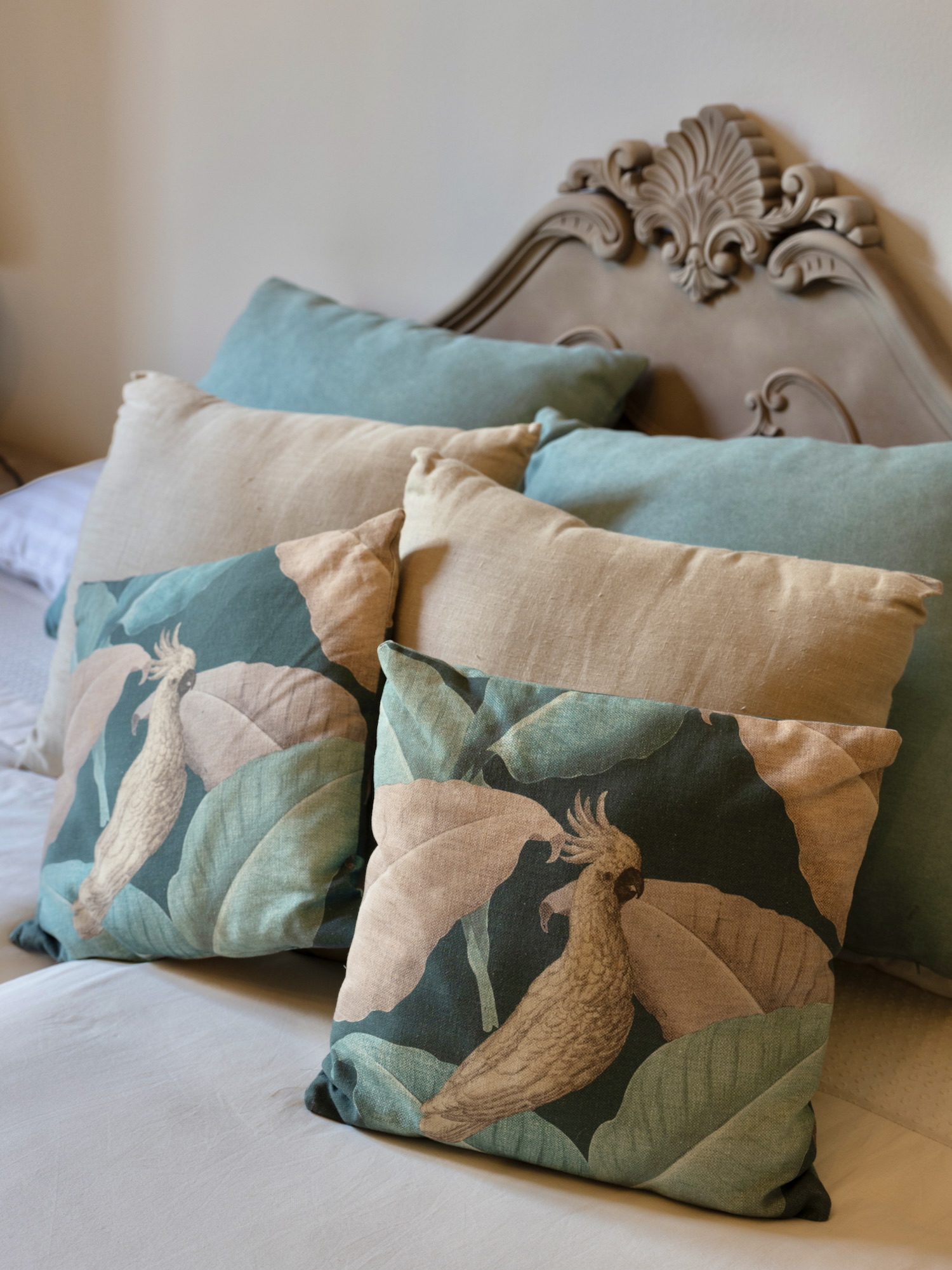Conjunto de cojines decorativos en tonos beige y verde agua con estampado de cacatúas y hojas tropicales, sobre una cama con cabecero ornamentado de estilo vintage