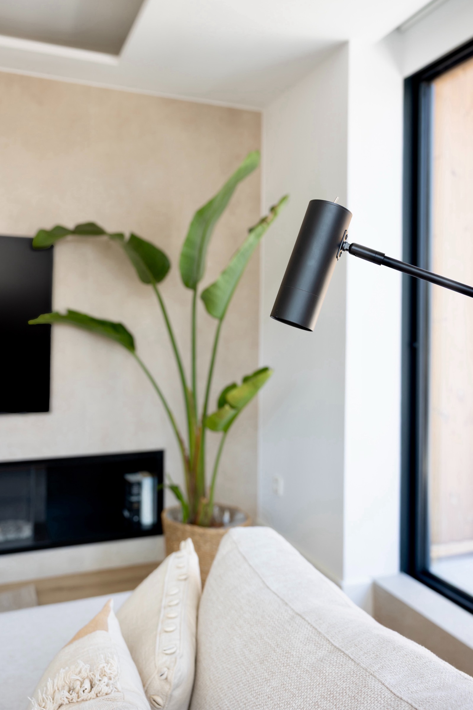 Interior de una sala moderna y minimalista con sofá beige, lámpara de brazo negra, planta grande en maceta y ventana grande. La decoración es limpia y luminosa con una pared de acento beige y una televisión montada en la pared.
