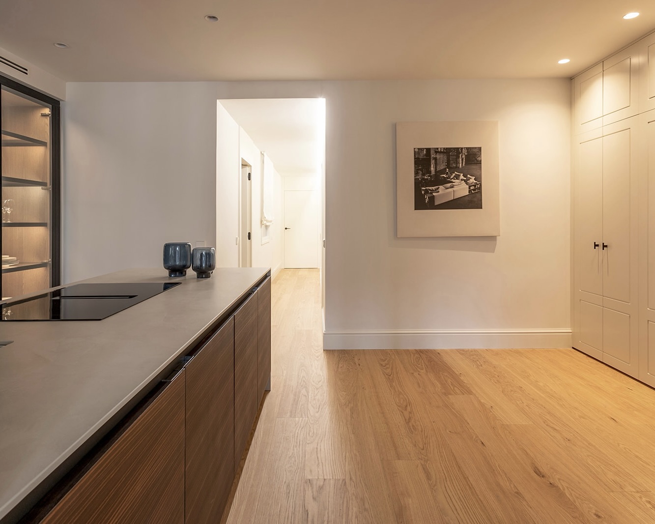 Cocina moderna y minimalista con isla de madera oscura, encimera gris, suelo de madera clara, armarios blancos empotrados y una obra de arte en blanco y negro en la pared. La iluminación es cálida y hay dos jarrones azules sobre la isla.