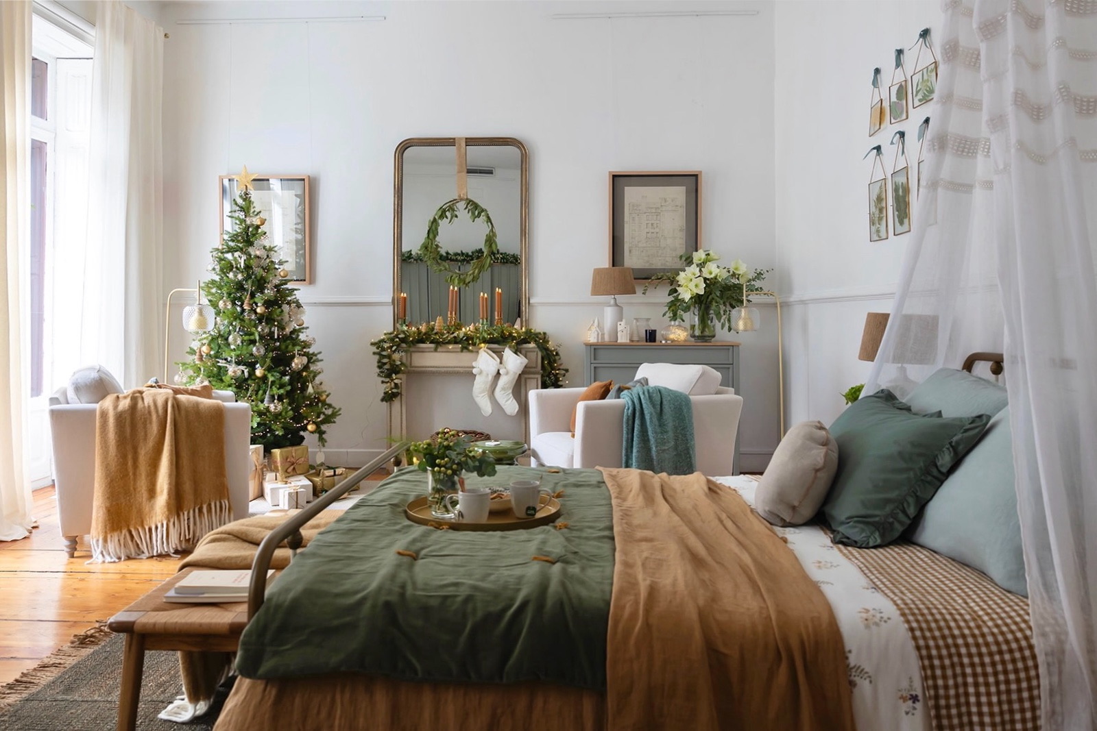 Dormitorio decorado para Navidad con árbol adornado, corona sobre espejo, velas y medias navideñas. Cama vestida en tonos verde y beige, con sillón blanco y detalles decorativos en estilo rústico moderno.