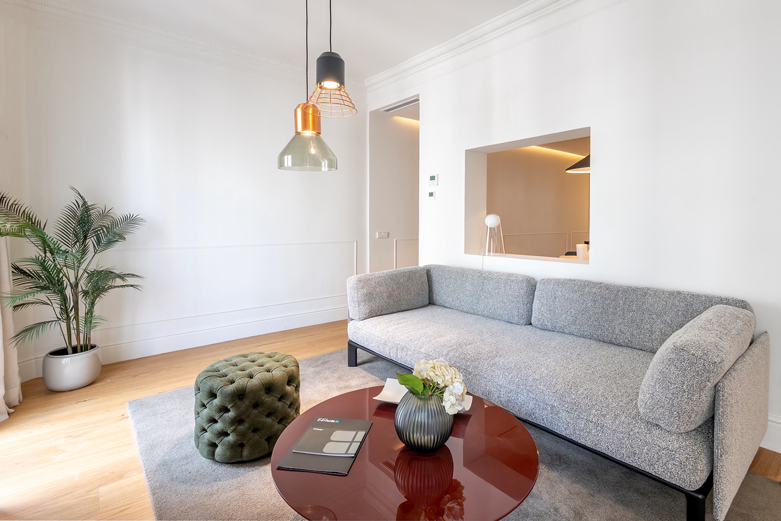 Sala de estar moderna y minimalista con sofá gris, mesa de centro roja, lámparas colgantes y planta decorativa. Espacio luminoso con paredes blancas y piso de madera.