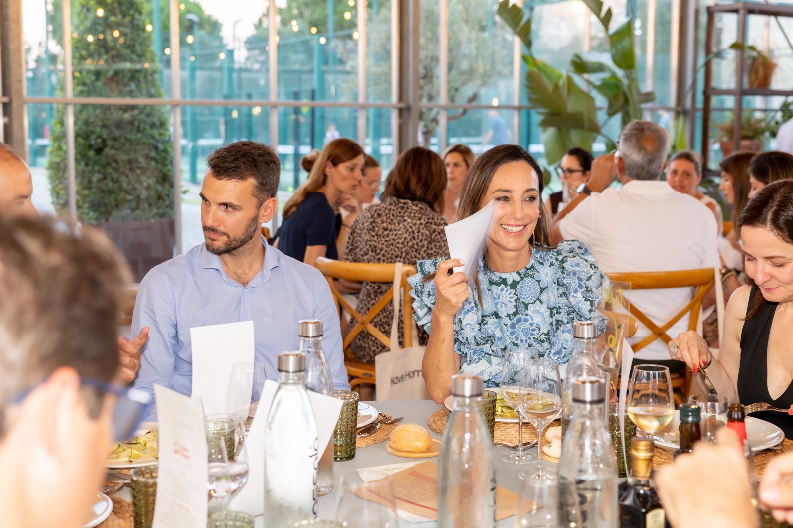 Grupo de personas sentadas en un evento social, almorzando en un espacio luminoso con grandes ventanales y plantas