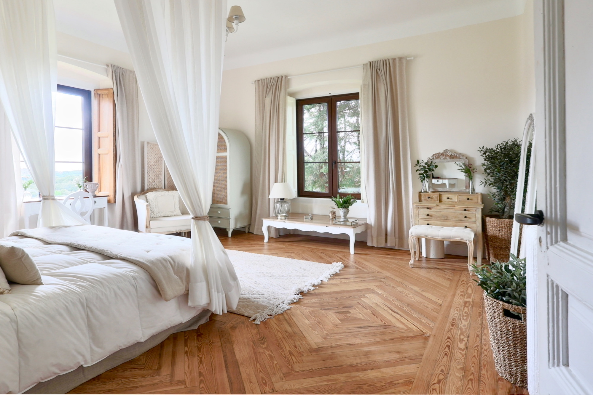 Dormitorio elegante en tonos blancos con piso de madera en espiga, cama con dosel, muebles estilo provenzal, cortinas beige y detalles decorativos en verde con plantas naturales. Ventanas grandes de madera permiten entrada de luz natural.