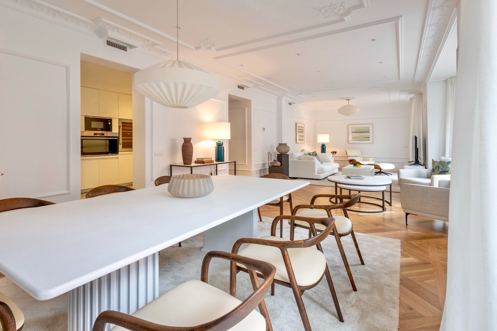 Interior de un apartamento moderno y elegante con comedor y sala de estar integrados. Mesa de comedor blanca con sillas de madera, lámparas colgantes decorativas, y una sala de estar con sofás blancos al fondo.