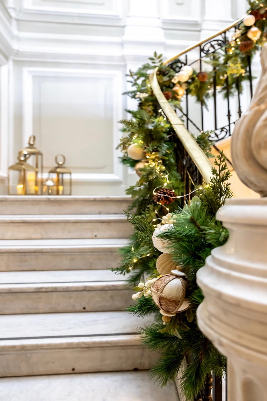 Elegante decoración navideña en escalera de mármol con guirnalda verde natural adornada con luces, bolas doradas y flores blancas. Faroles dorados iluminados en el fondo y barandilla de hierro forjado negro