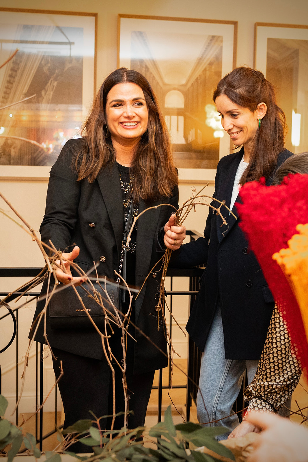 Dos mujeres sonrientes arreglando ramas secas en un evento. Visten abrigos negros y están rodeadas de flores y decoraciones. Al fondo se ven cuadros enmarcados en la pared.