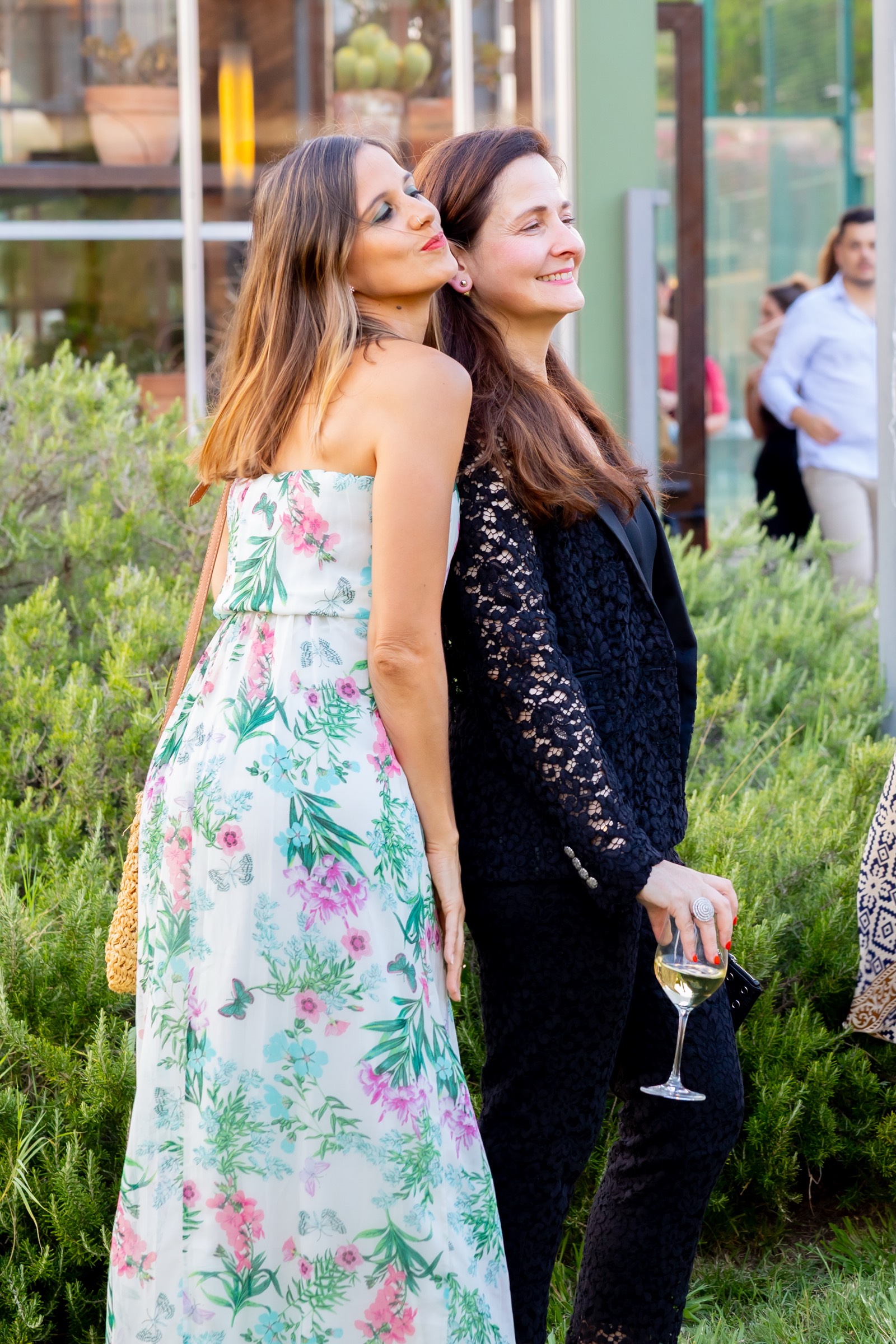Dos mujeres de pie en un jardín, una con vestido floral blanco y verde, la otra con vestido negro de encaje, sosteniendo una copa de vino