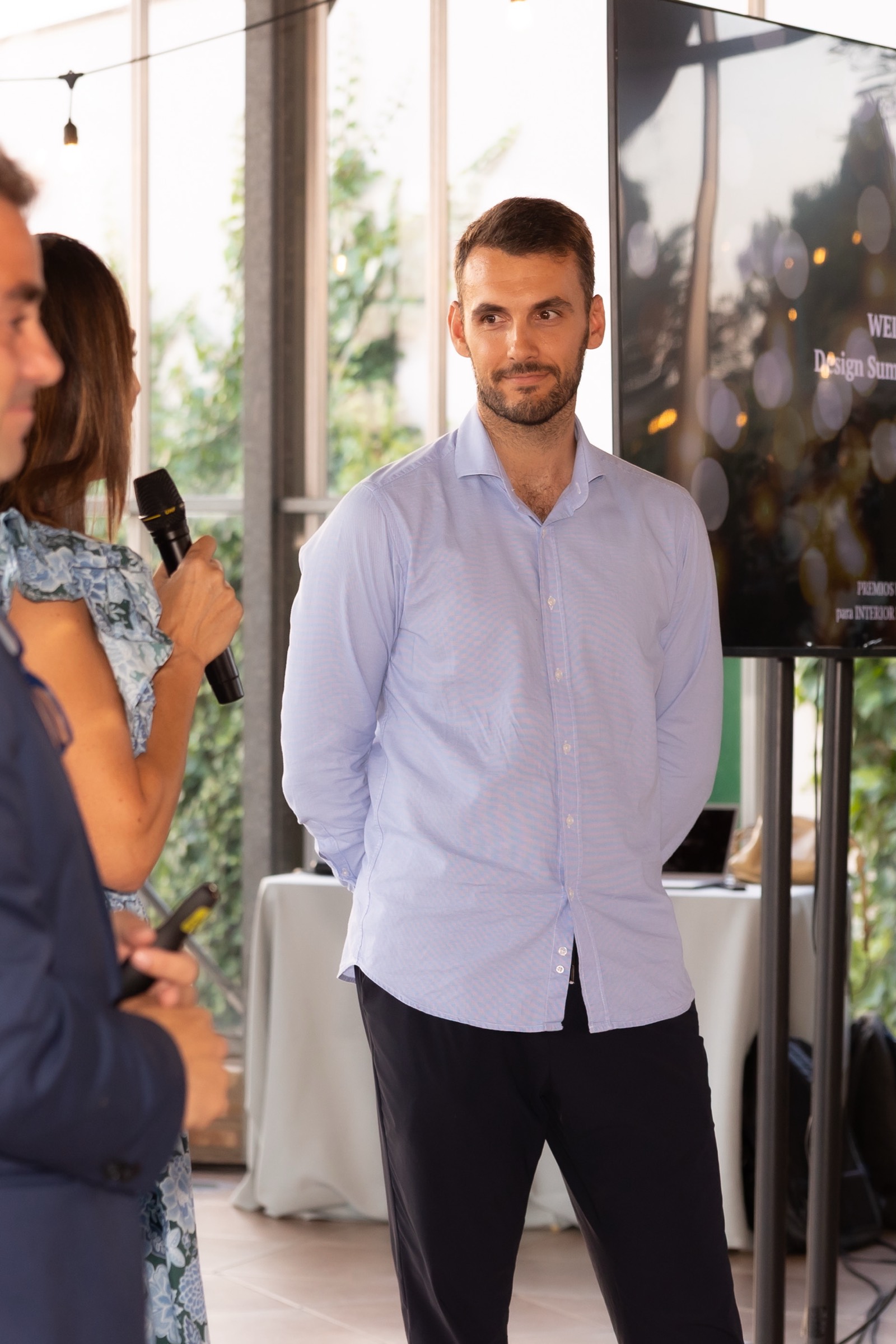 Escena de evento corporativo con participantes en un espacio moderno con grandes ventanales, uno de ellos sosteniendo un micrófono