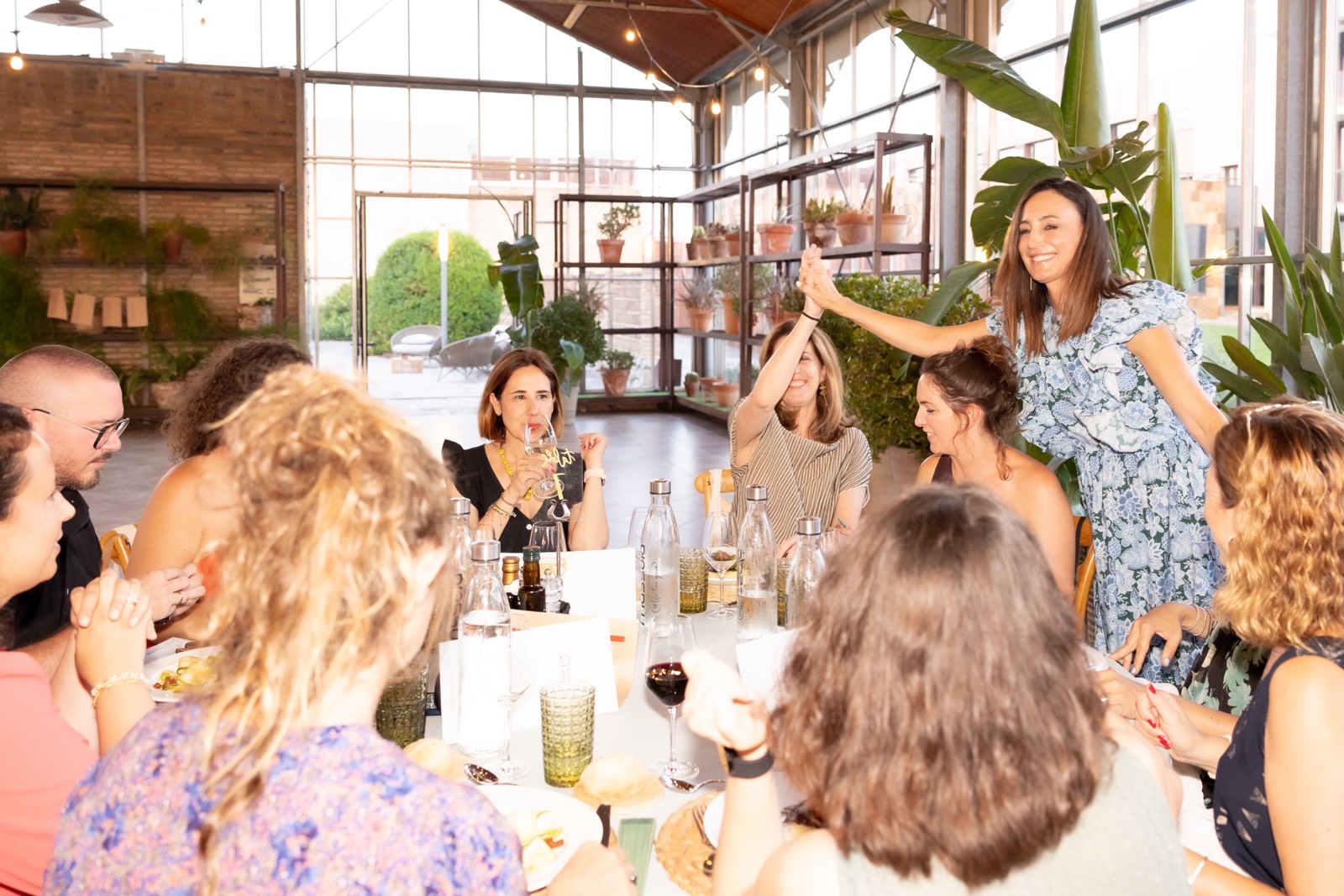 Grupo de mujeres reunidas alrededor de una mesa en un restaurante con plantas y luz natural, compartiendo bebidas y conversación