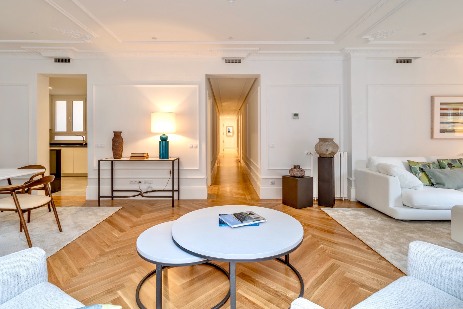 Sala de estar elegante y moderna con paredes blancas, piso de madera en espiga, muebles blancos, mesas de centro redondas y decoración minimalista. Un pasillo iluminado conduce a otras áreas de la casa.