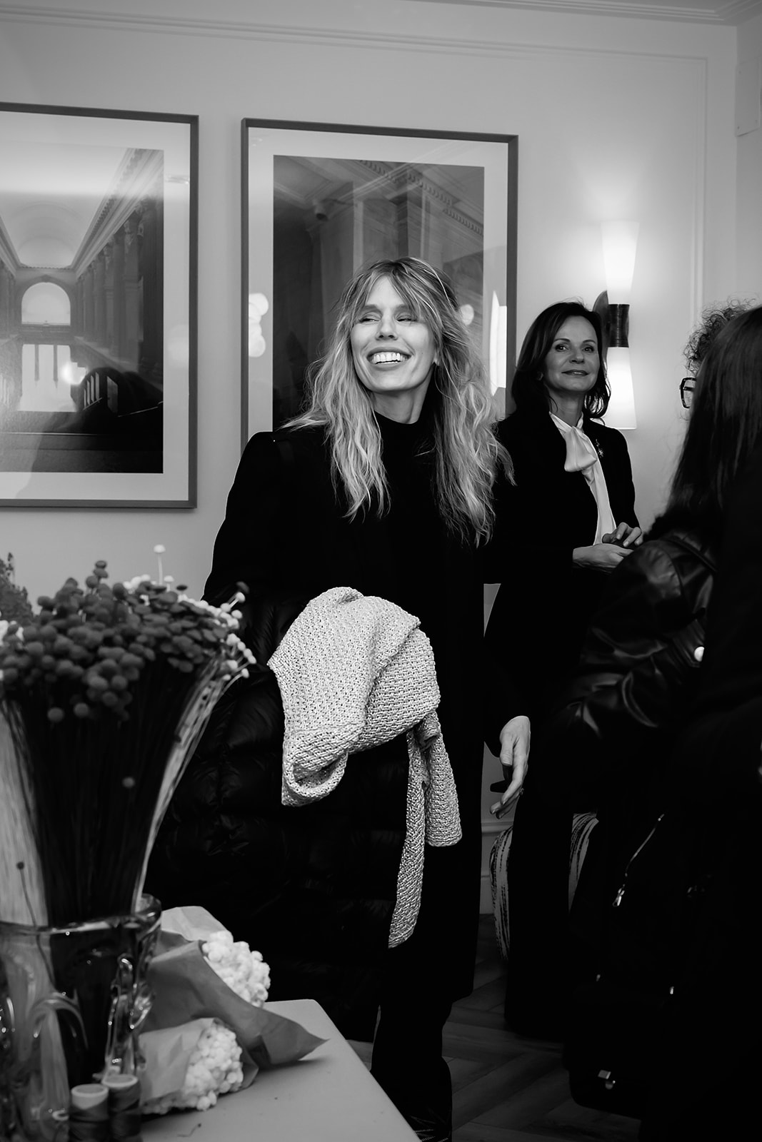 Imagen en blanco y negro de mujeres sonrientes en un evento elegante. Una mujer rubia en primer plano sonríe ampliamente, sosteniendo una prenda blanca. Detrás, otra mujer sonríe. Hay cuadros en la pared y un arreglo floral visible.