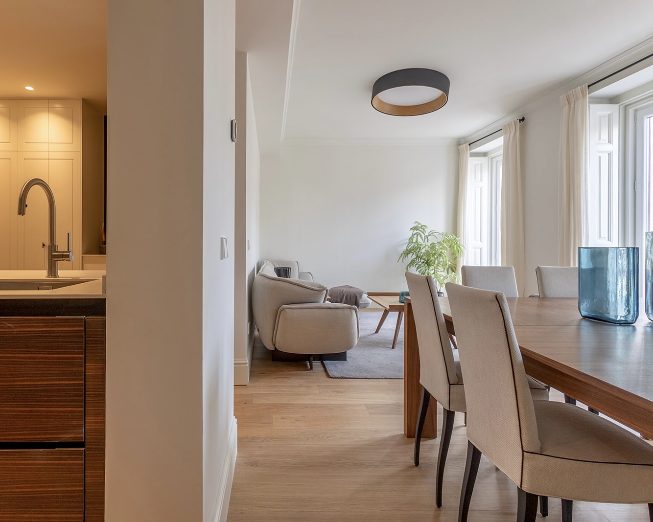 Interior de un apartamento moderno con cocina abierta, comedor y sala de estar. Decoración minimalista con muebles en tonos neutros, pisos de madera clara y grandes ventanas que permiten la entrada de luz natural.
